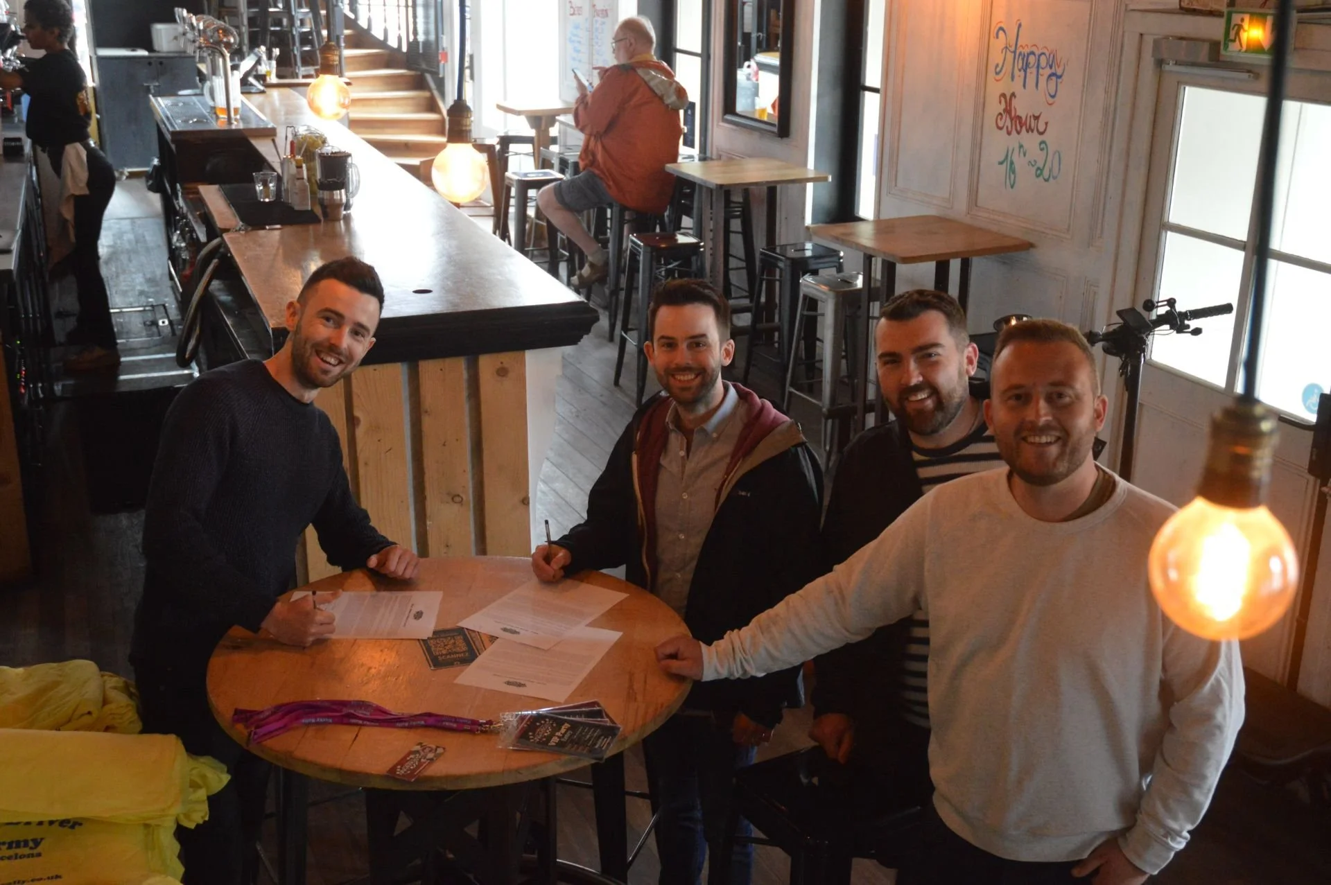 Four men smiling and signing papers around a round wooden table in a cozy cafe or restaurant with wooden floors and wall decor.