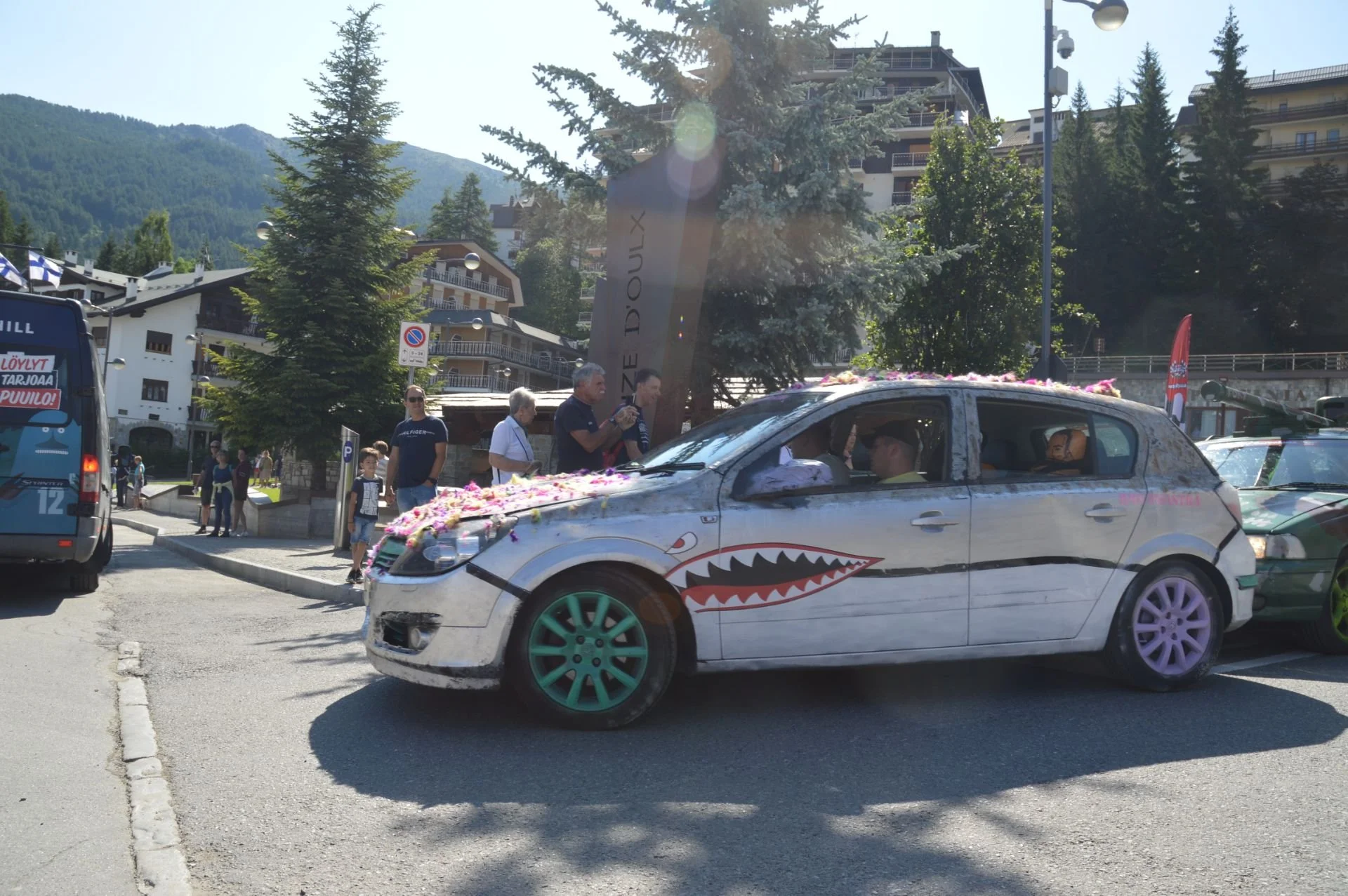 A decorated car with shark mouth design, colorful wheels, and flower arrangements on top, parked on a street with people, trees, mountains, and buildings in the background.