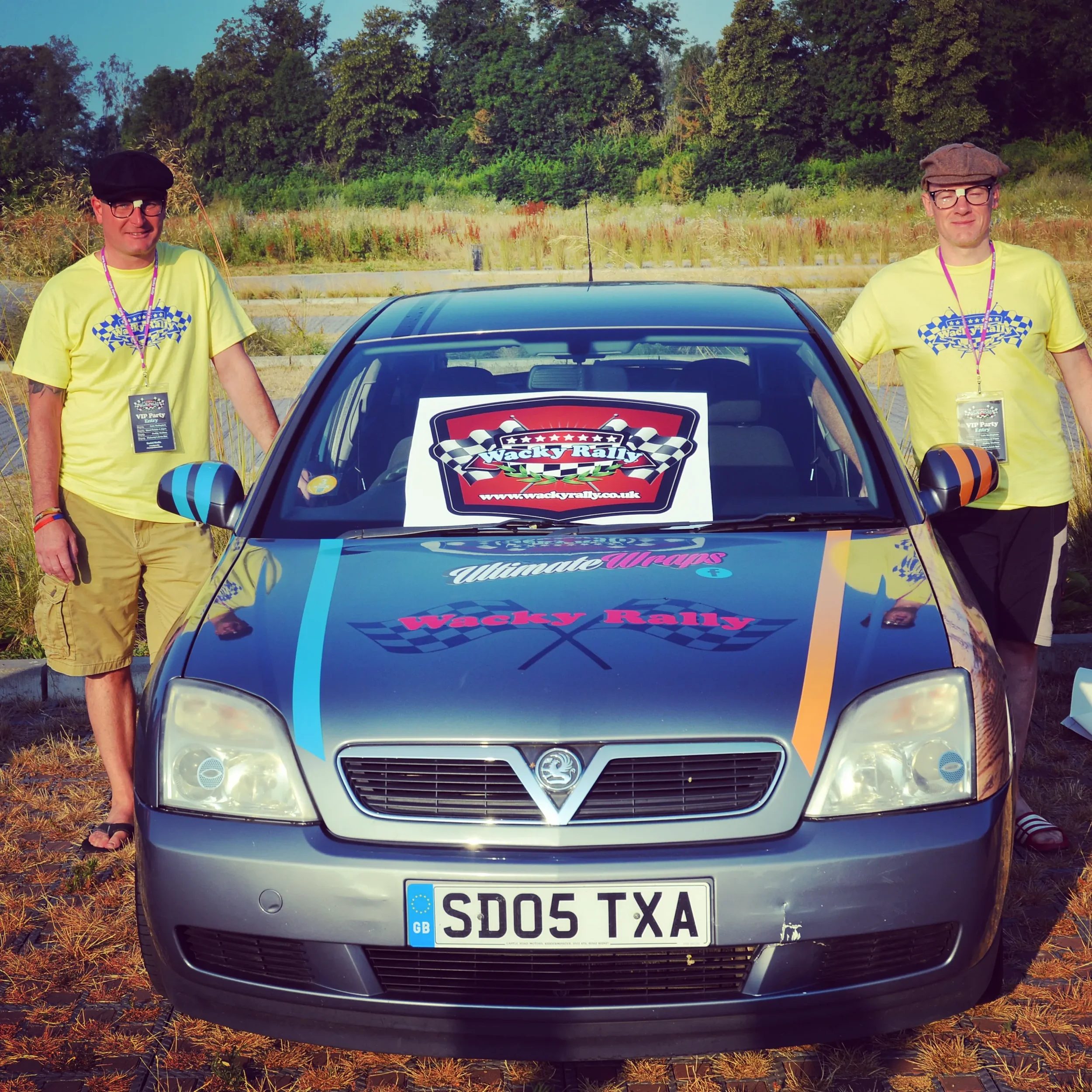 Two men standing next to a silver Vauxhall car with rally-themed decals, including the words "Wacky Rally" and a sign on the windshield, in a grassy outdoor area with trees in the background.