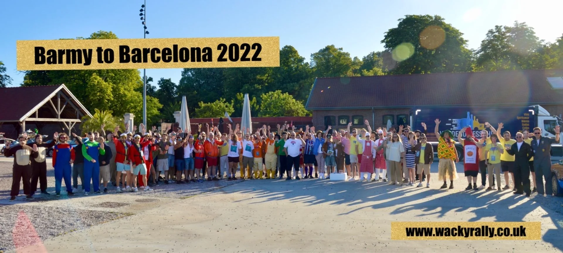 Large group of people in costumes and casual clothing standing outdoors, raising their hands, with trees and buildings in the background during daytime. Text overlay reads 'Barmy to Barcelona 2022' and the URL 'www.wackyrally.co.uk'.