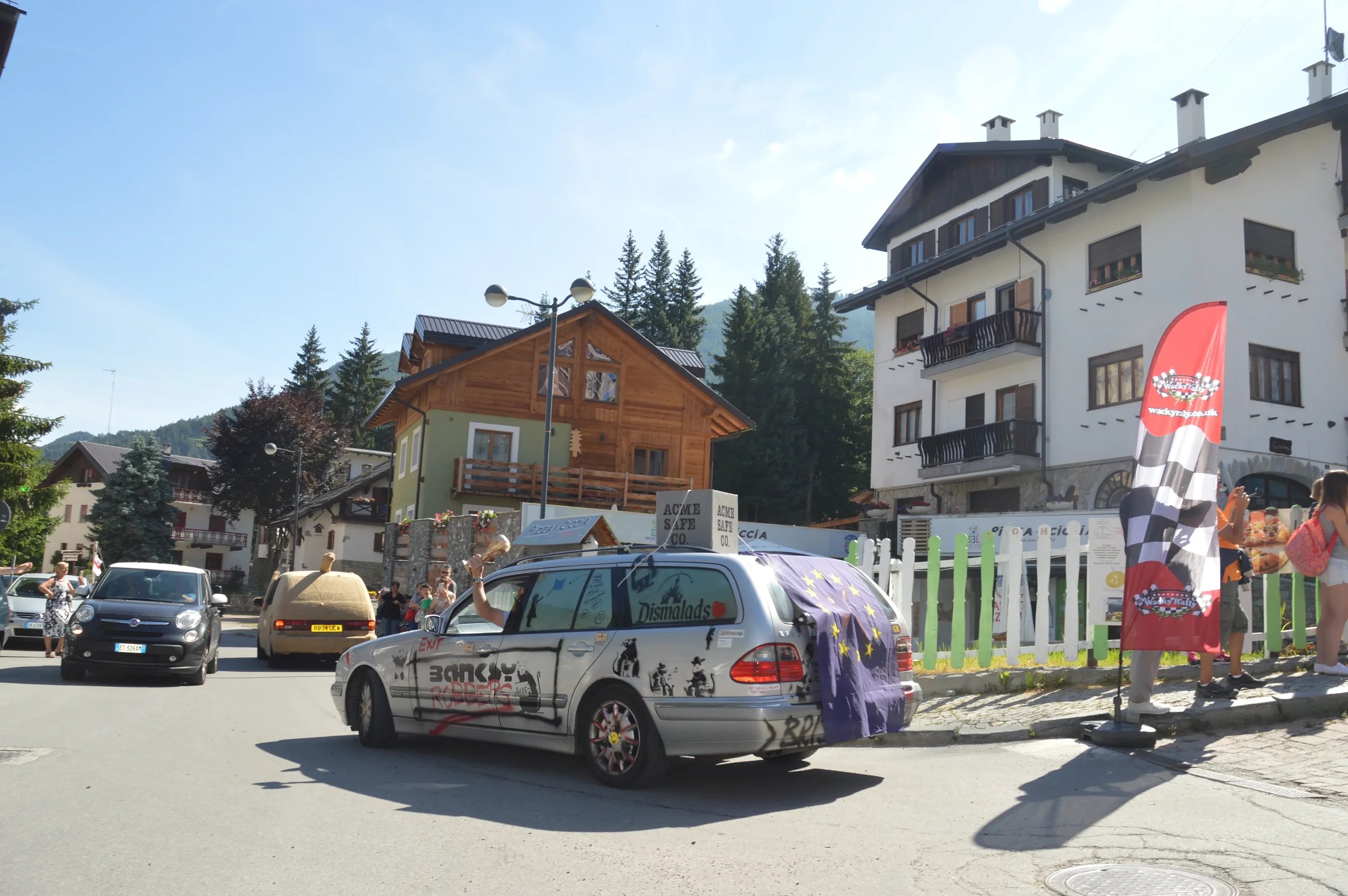 Vehicles parked on a street in a mountain village with buildings and trees in the background, some people standing near the cars, and a flag with a racing checkered pattern.