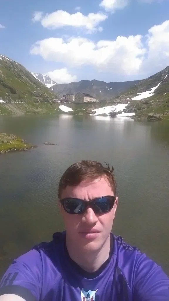 Young man wearing sunglasses and a purple sports shirt taking a selfie near a mountain lake with snow patches, hills, and buildings in the background under a partly cloudy sky.