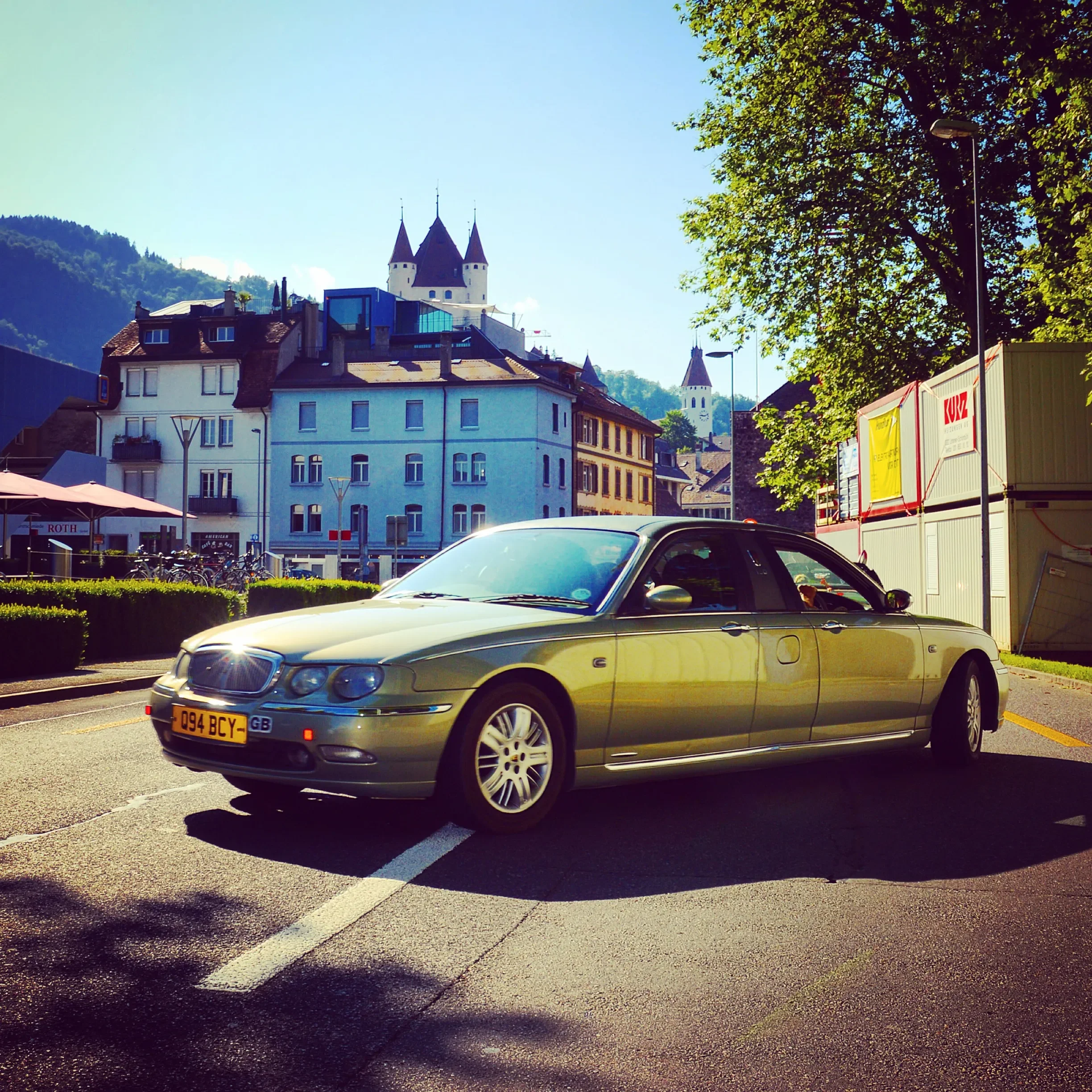 A yellow sedan parked on a city street with buildings and a castle in the background, sunny weather, and a large tree casting shadows.