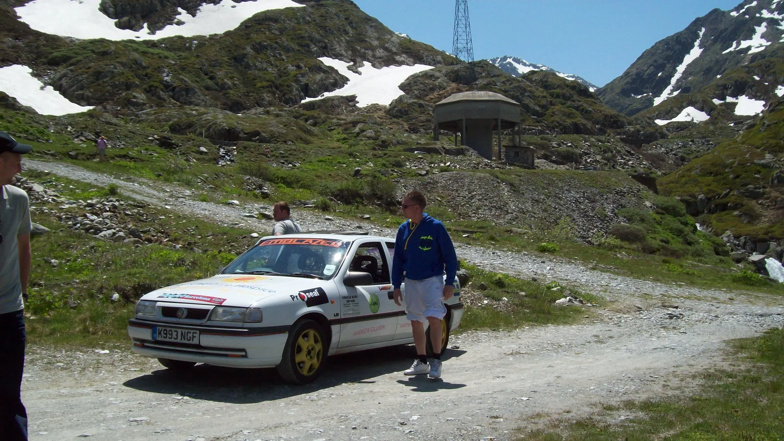 A group of people near a rally car on a dirt mountain trail with snowy peaks and green hills in the background.
