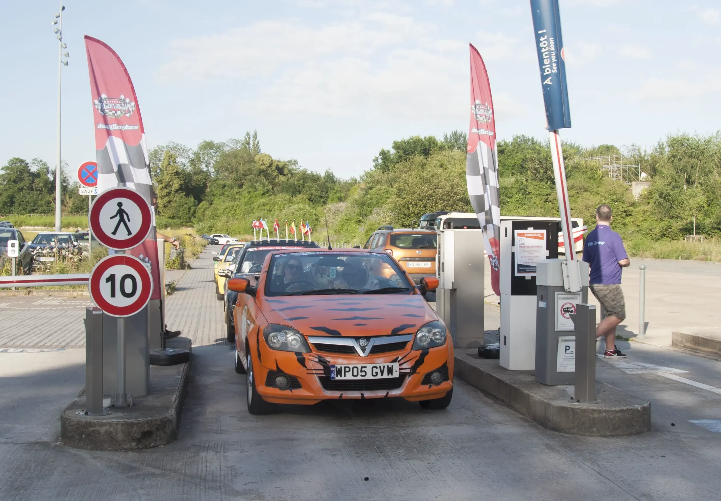 Orange car with black tiger stripe pattern at parking lot gate, man walking nearby, flags in the background, parking signs, and a row of cars parked.