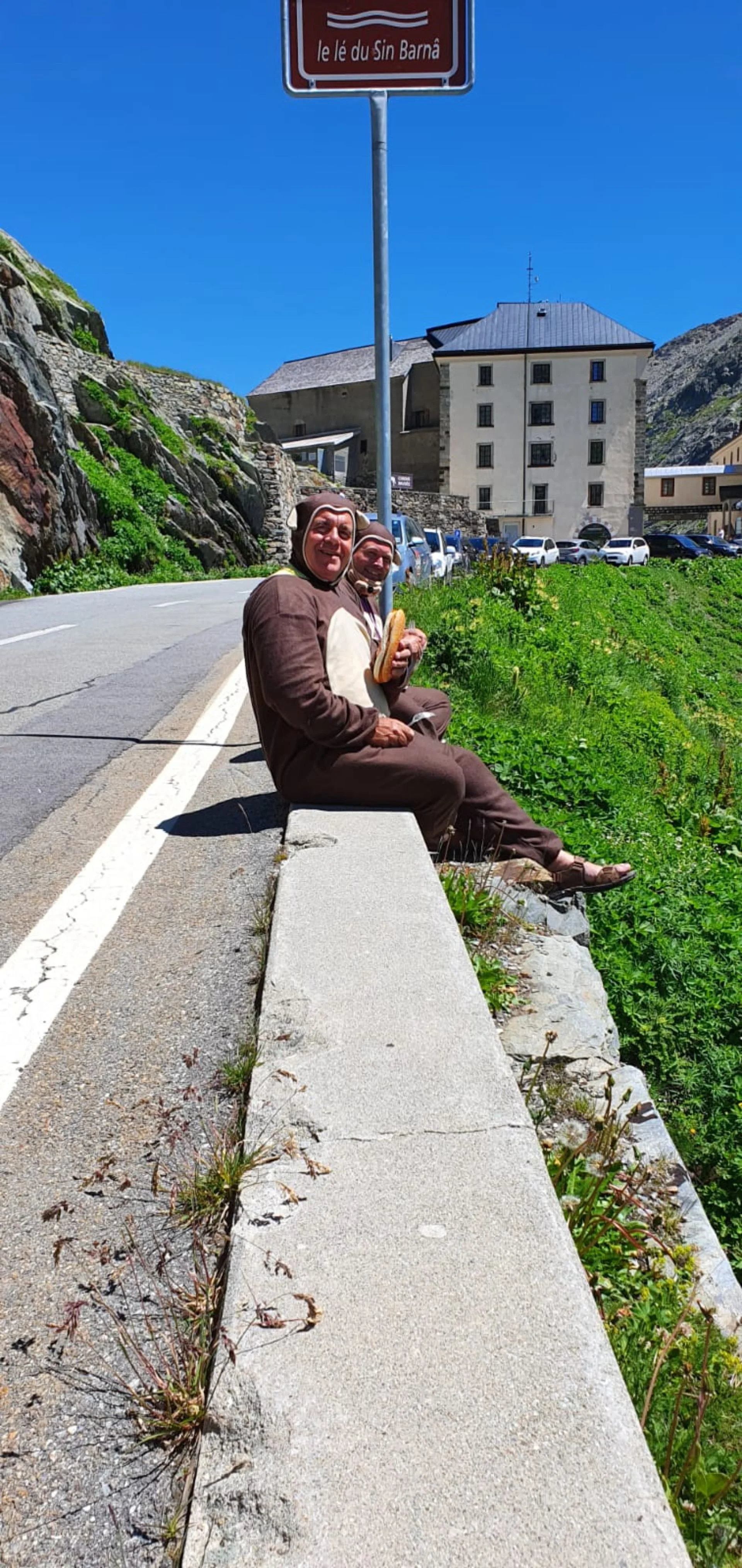 Two people dressed in dog costumes sitting on a bench by the roadside, holding baguettes, with a building, parked cars, and a signpost in the background on a sunny day.