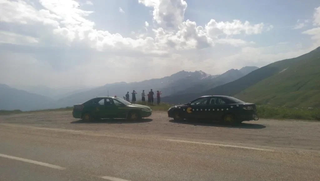 Two police cars parked on a mountain road with a group of people standing on the side, scenic mountain landscape in the background.