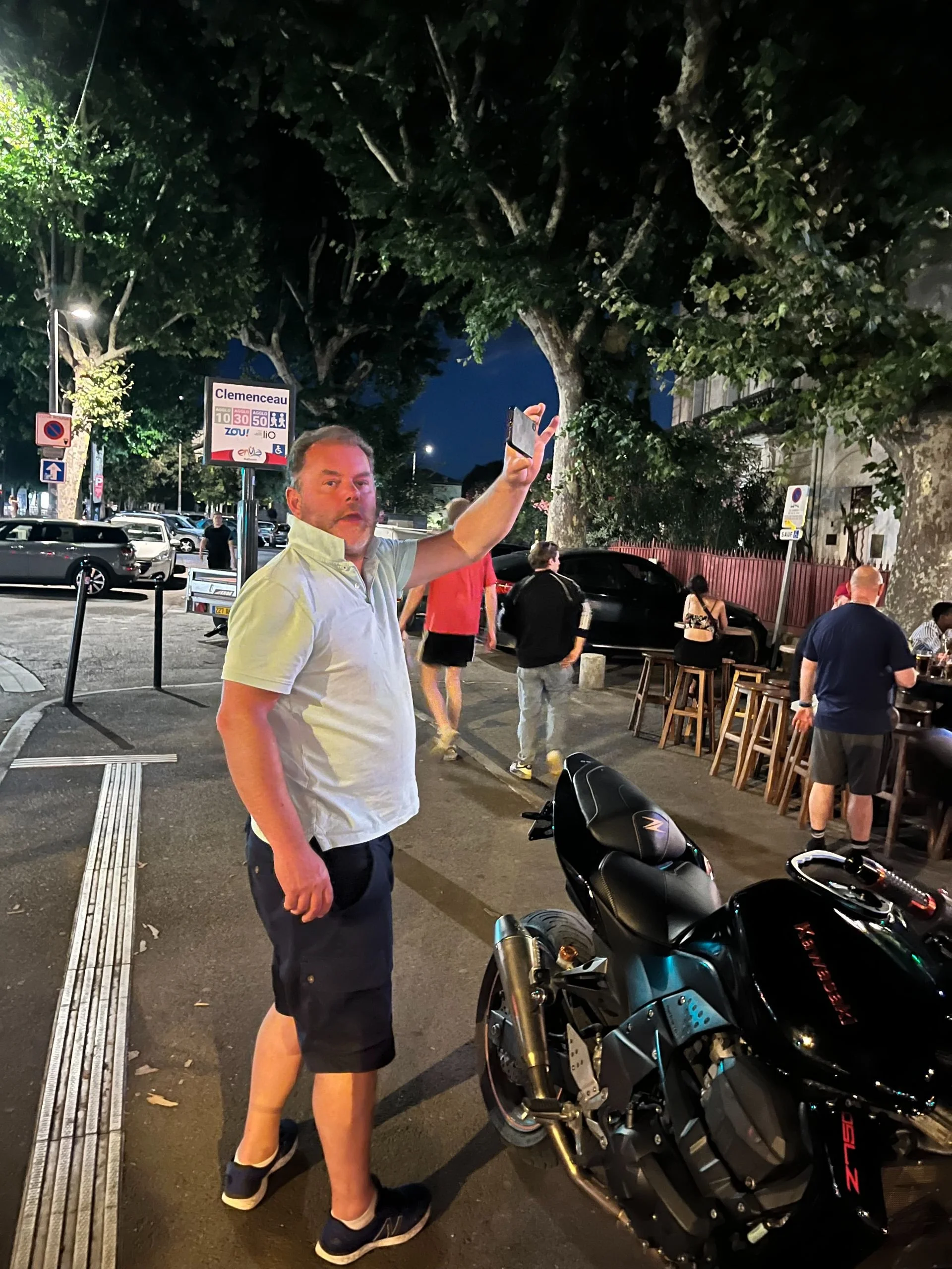 A man in a white polo shirt and black shorts takes a selfie at night in an outdoor setting with trees, parked cars, and people sitting and standing near a bar or restaurant. A black motorcycle is parked nearby.
