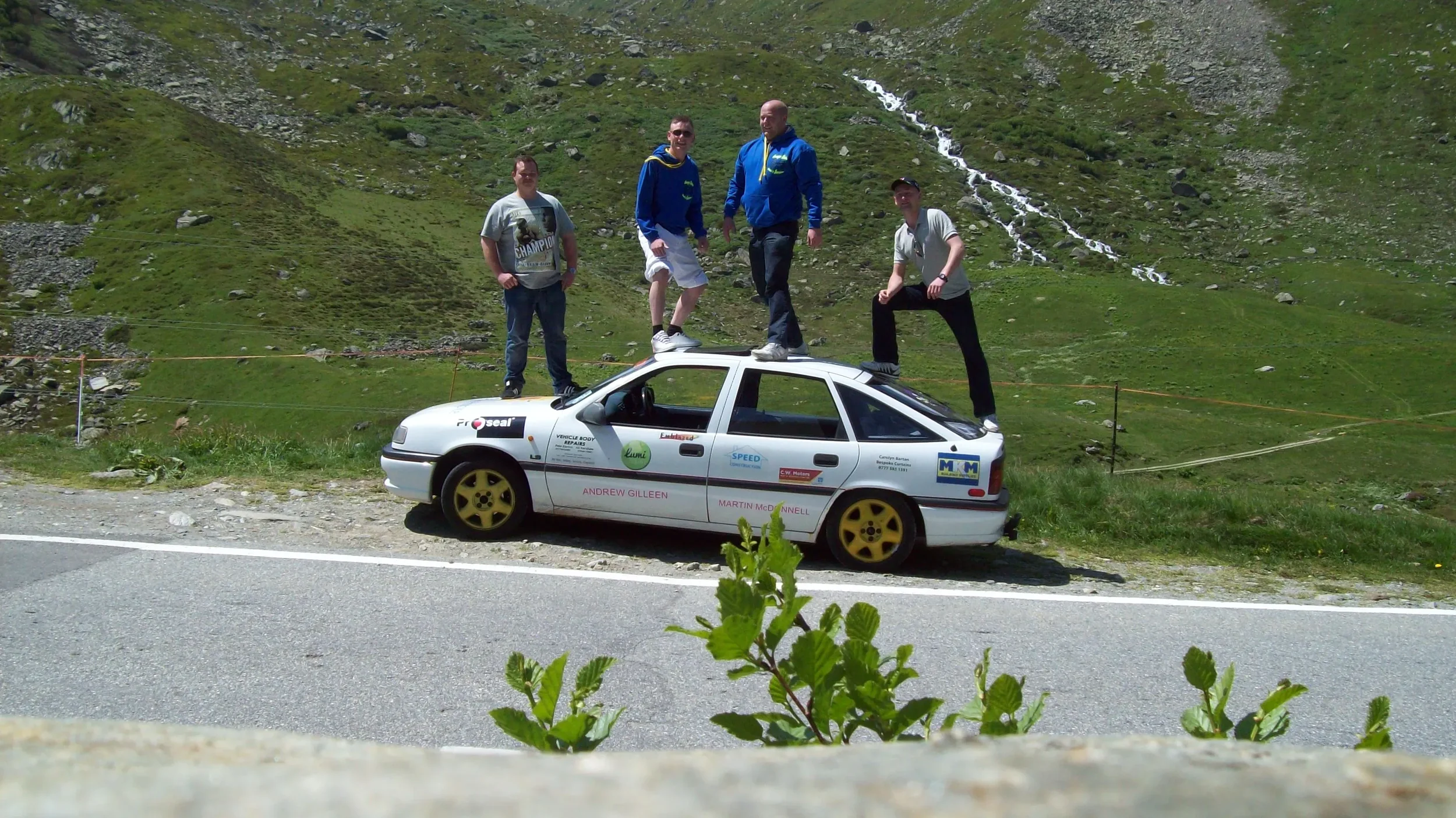 Four men standing and kneeling on and around a modified white race car with yellow wheels on a grassy mountain road, with a green mountain landscape and small waterfalls in the background.