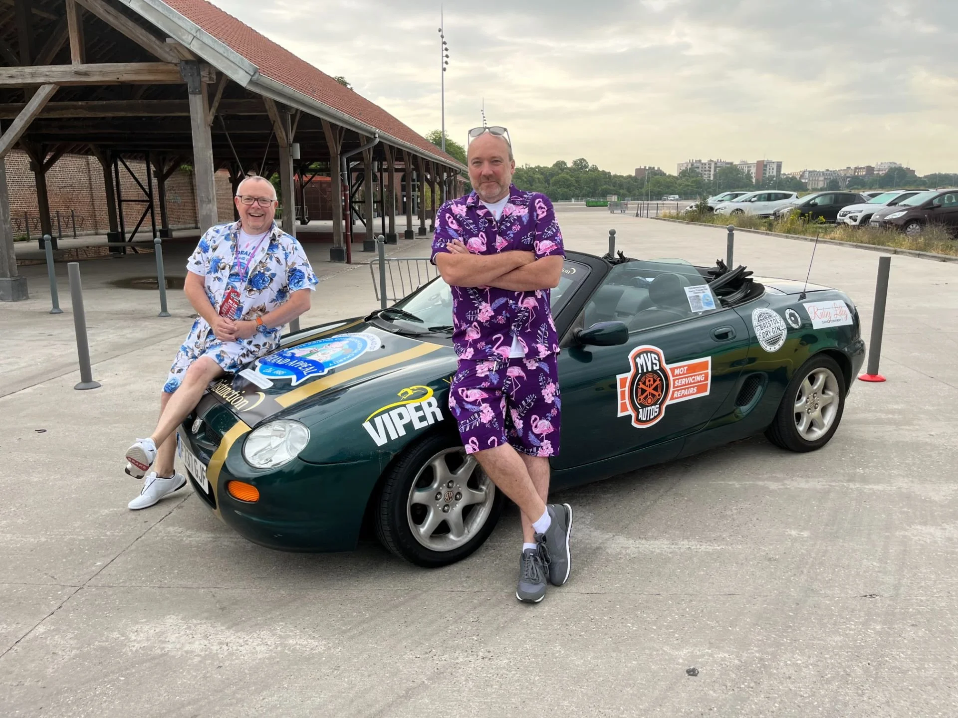 Two men in colorful Hawaiian shirts and shorts leaning against a vintage convertible sports car in a parking lot. One is sitting on the car's hood smiling, and the other is standing with arms crossed. The car has various stickers and sponsor decals.
