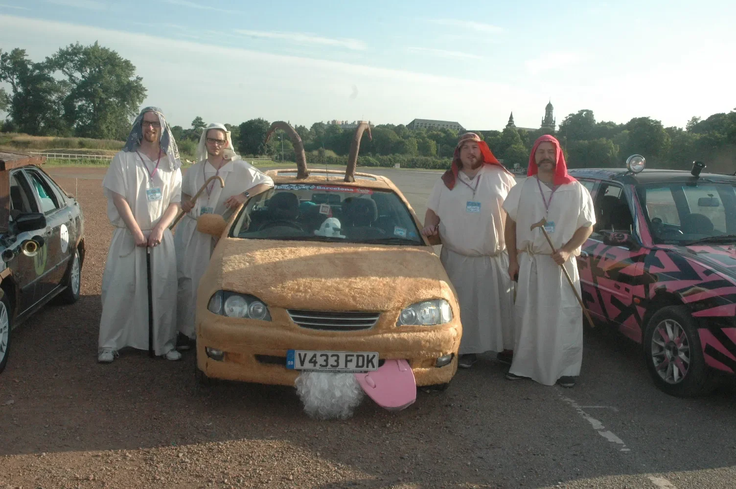 Group of five people dressed as biblical characters standing around a car covered in gold fabric, with two of them holding tools and one with a basket, in a parking lot with other decorated cars.