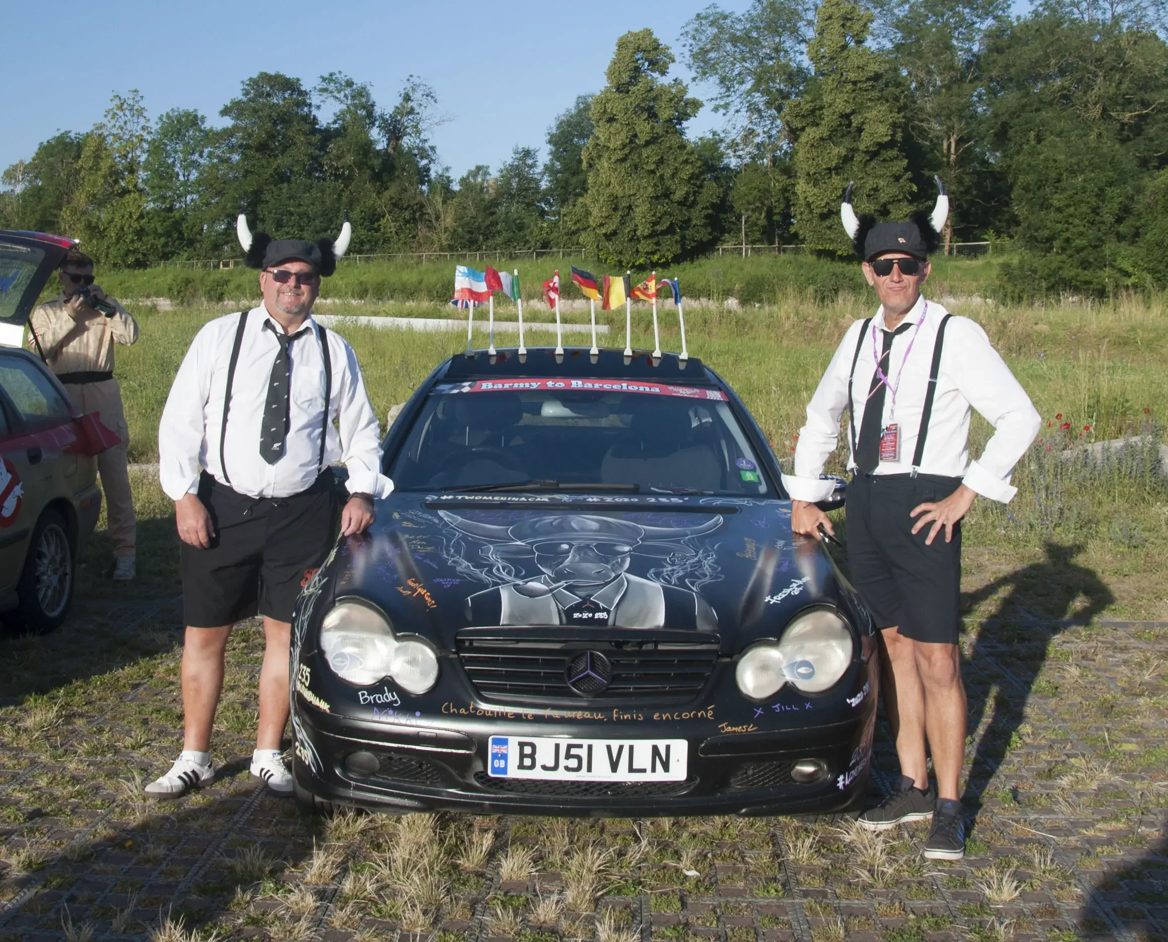 Two men dressed in white shirts, black shorts, suspenders, and Viking helmets with horns, standing next to a black Mercedes-Benz sports car decorated with signatures and drawings, with flags on top, in an outdoor grassy area.