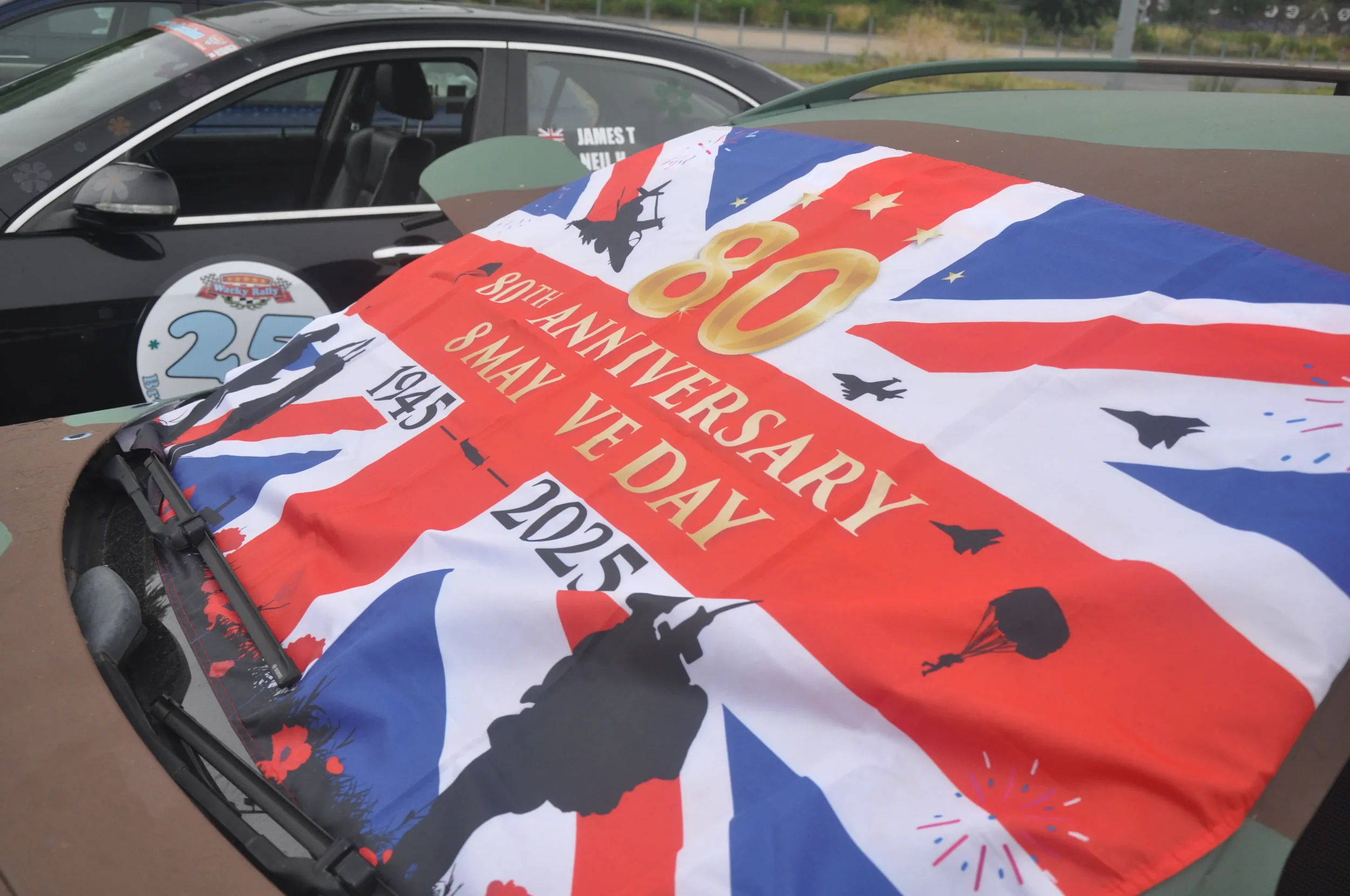 A large banner on a car celebrating the 80th anniversary of VE Day, with a Union Jack flag design, silhouettes of soldiers, parachutes, and aircraft, and text noting May 8, 1945, to May 8, 2025.