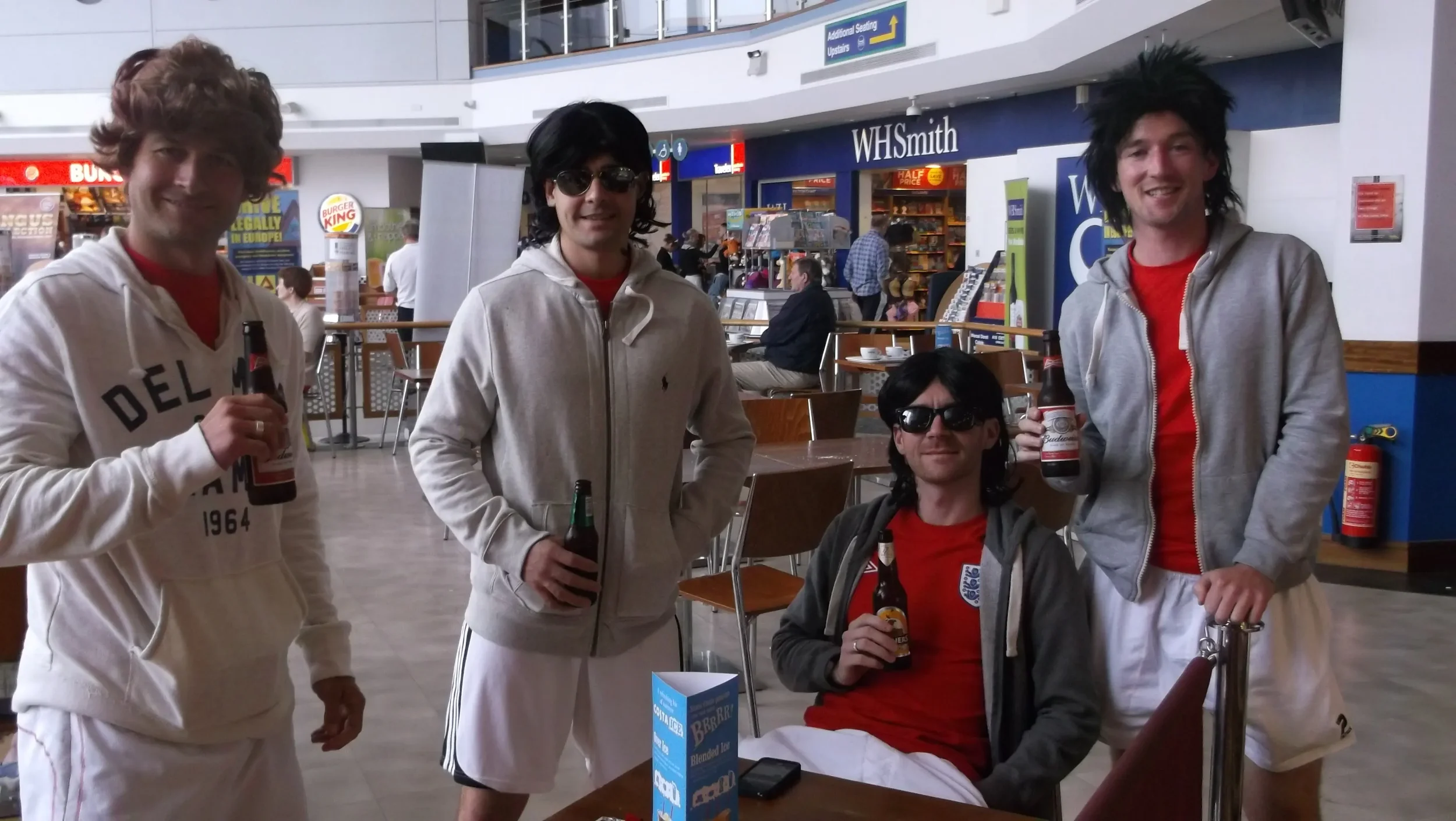 Four men dressed as Beatles in a mall, holding beer bottles, with a background of a food court and stores.
