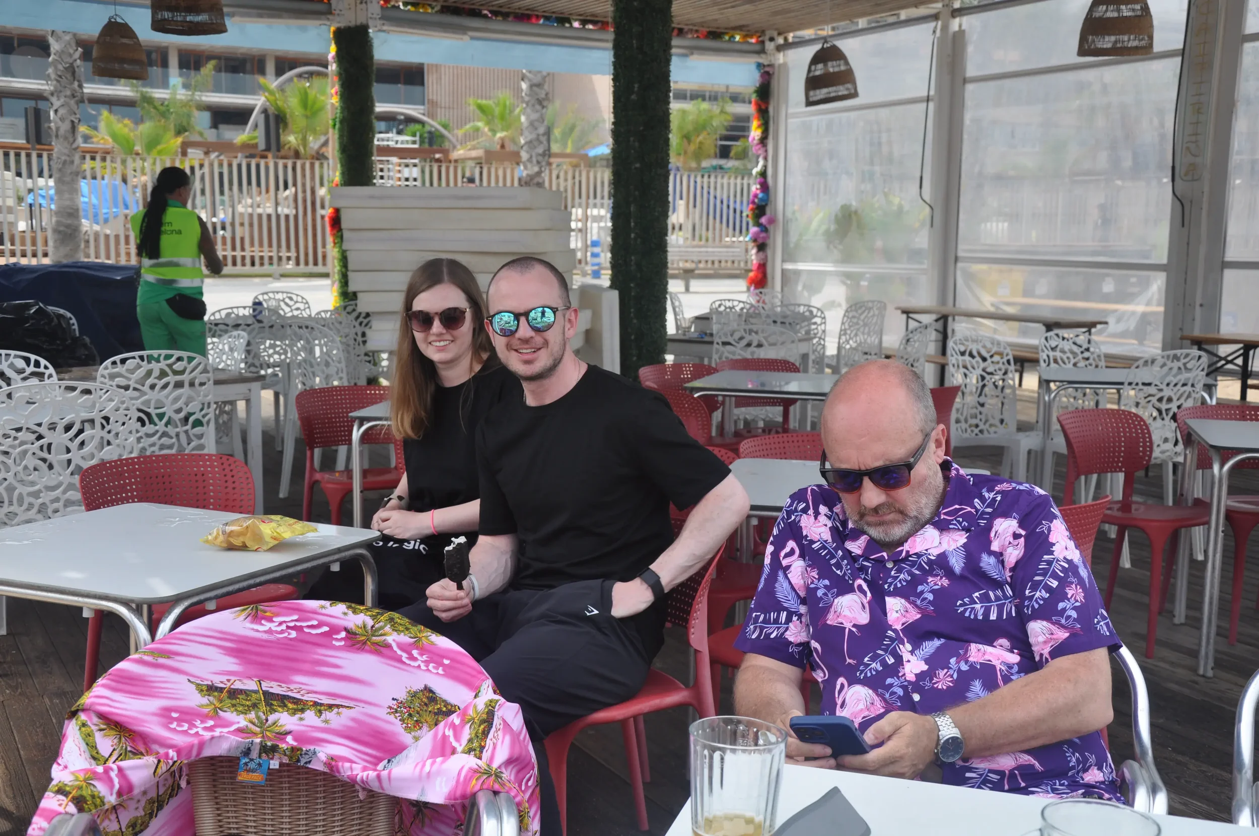 Three people sitting at outdoor table at a beachside restaurant, two men and one woman, wearing sunglasses, with palm trees and a person in green in the background.