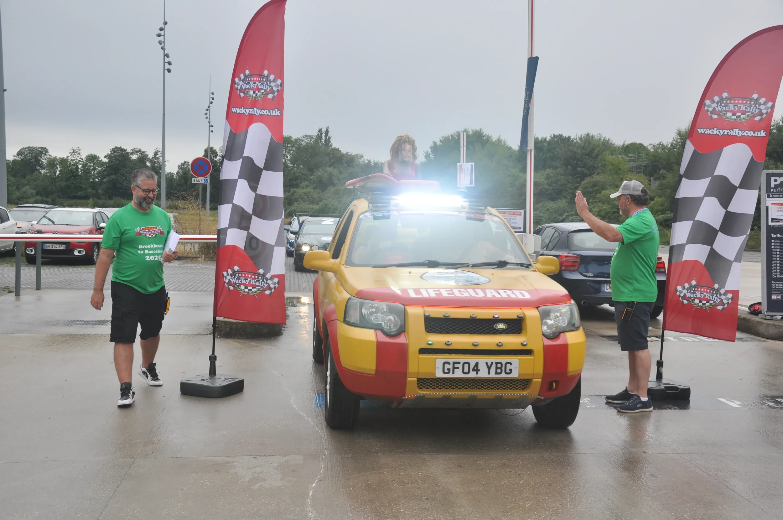 A yellow lifeguard vehicle at a rally event with two men in green shirts standing on either side, one waving and the other walking while holding papers, near checkered flag banners, on a wet parking lot.