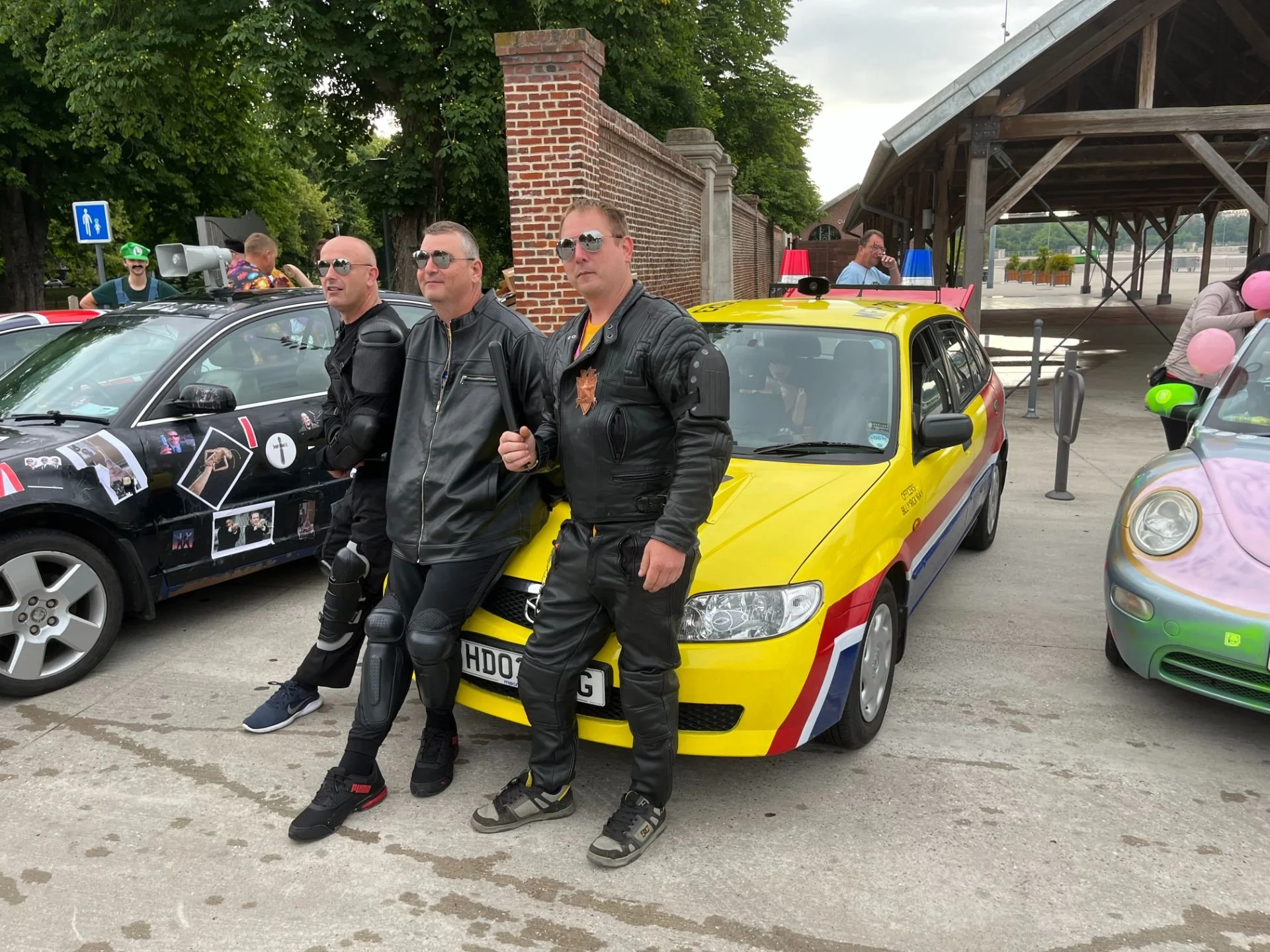 Three men in leather jackets and sunglasses posing in front of a yellow and red rally car at a car meet, with other decorated cars and people in the background.