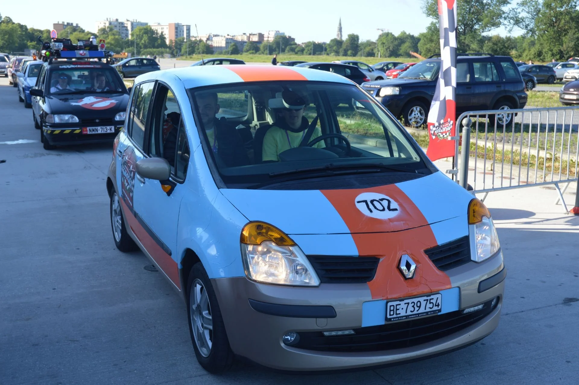 A Renault car with racing decals and the number 702 on the hood, part of a convoy with other car race vehicles in the background, at an outdoor event with cars parked nearby.