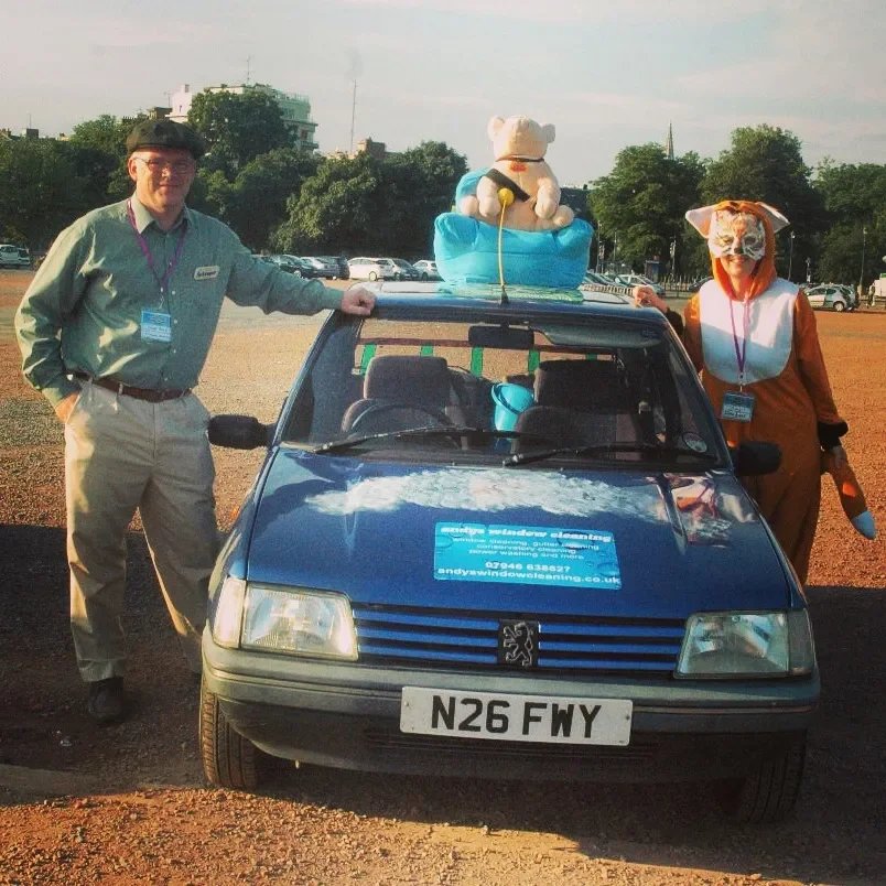 Two people in costumes standing next to a small blue car with a large stuffed bear on top and a sign on the hood, in a parking lot with trees and buildings in the background.