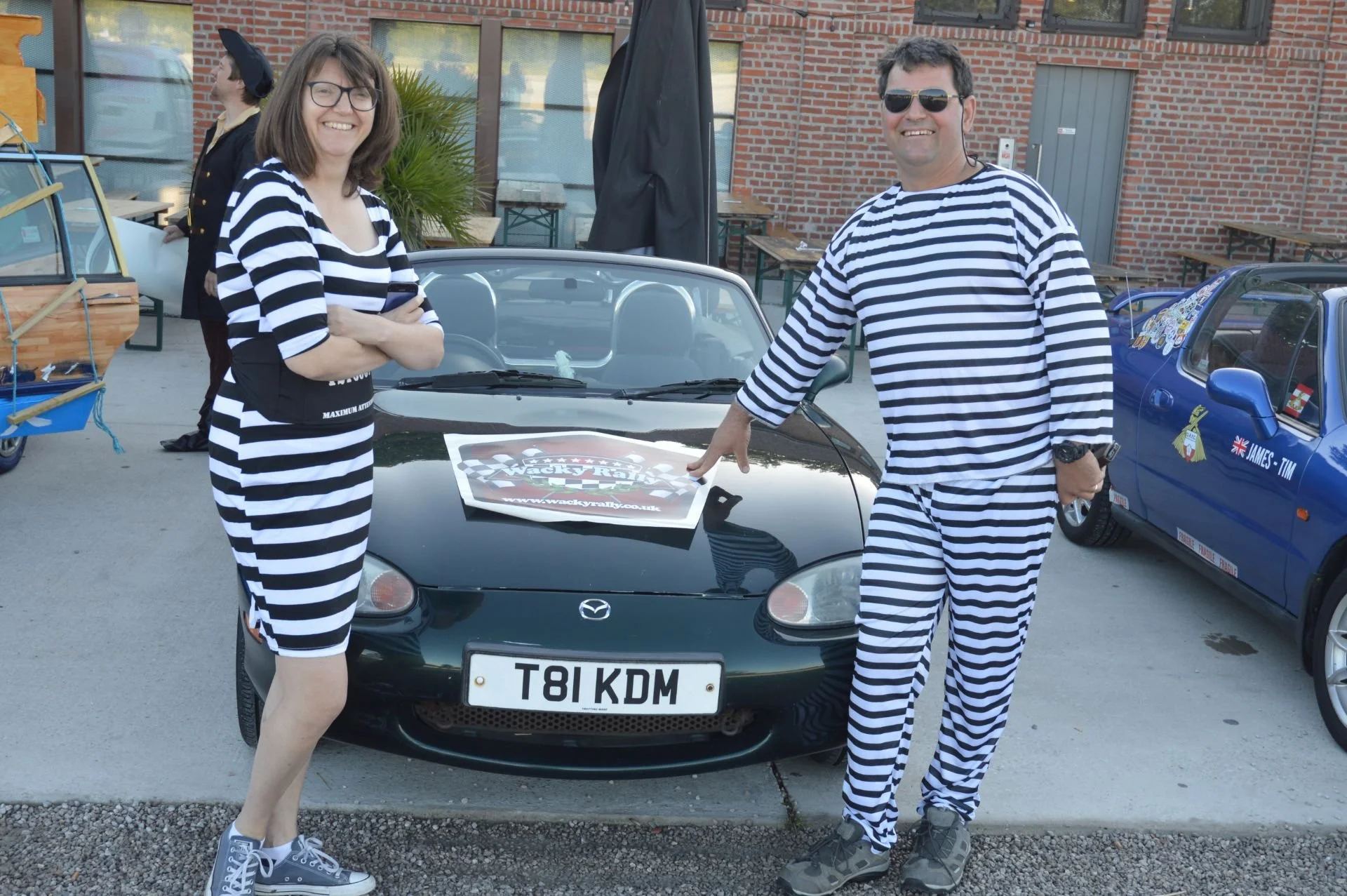 Two people dressed in matching black and white striped outfits standing in front of a black Mazda sports car at a car rally event. The woman on the left is smiling with arms crossed, while the man on the right is pointing to the car's hood with a smi
