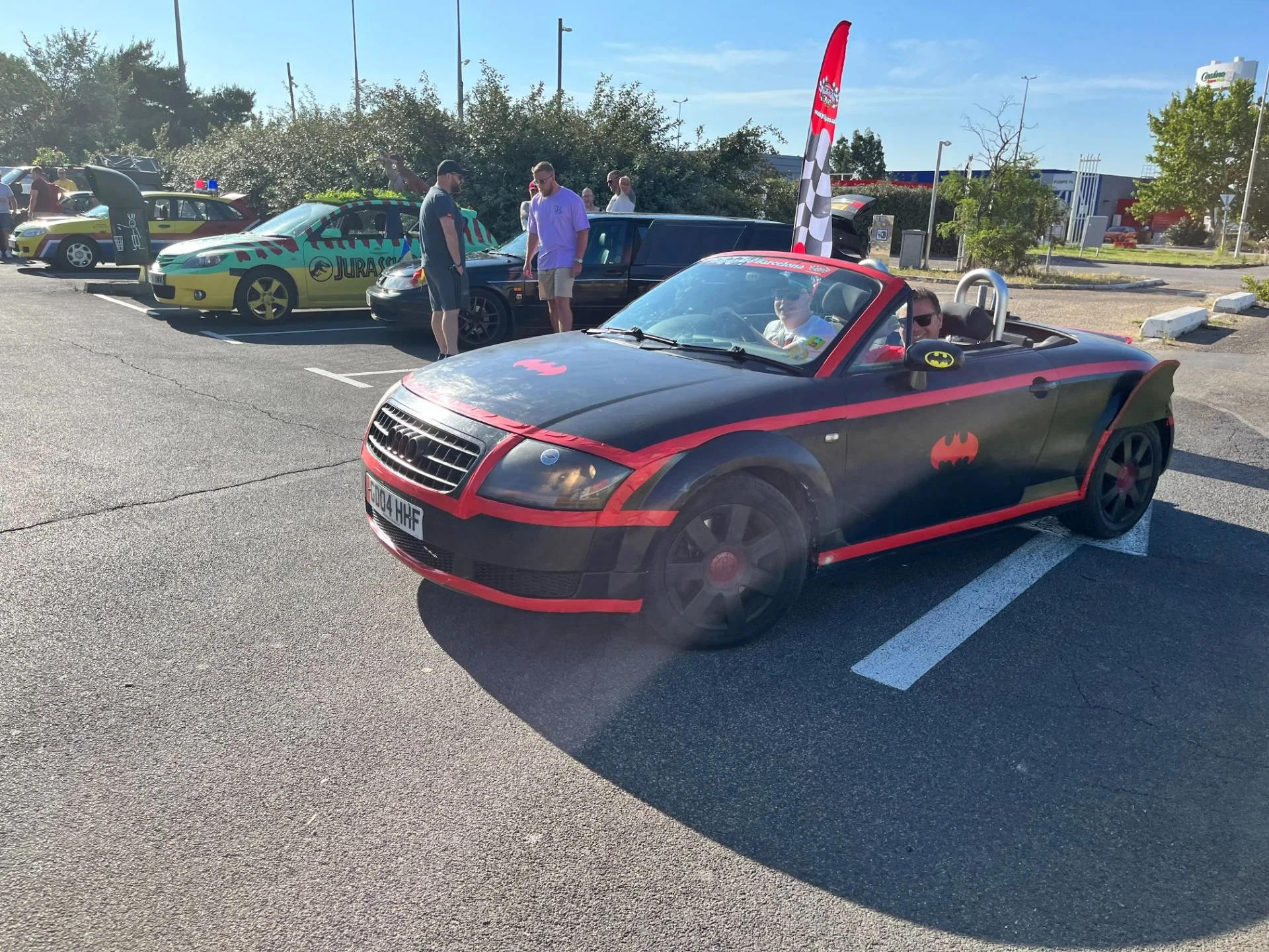 A black Audi sports car with Batman theme, parked in a parking lot during daylight. The car has Batman logos on the hood, door, and side mirrors, with red accents on the edges. Inside, two people are visible, with one wearing sunglasses. In the backg