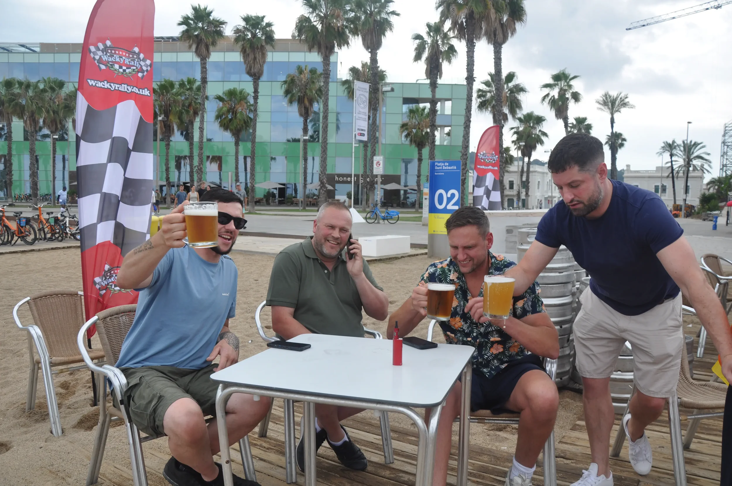 Four men sitting at a table on a beach, holding beers and smiling, with palm trees, modern buildings, and flags in the background.