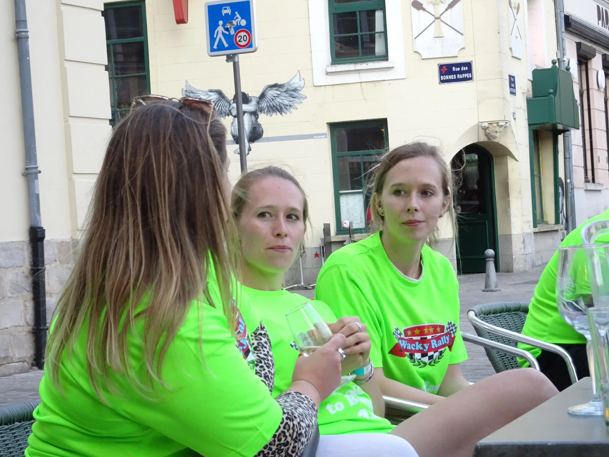Three young women in bright green T-shirts sitting at an outdoor table, holding glasses, with a building and street sign in the background.