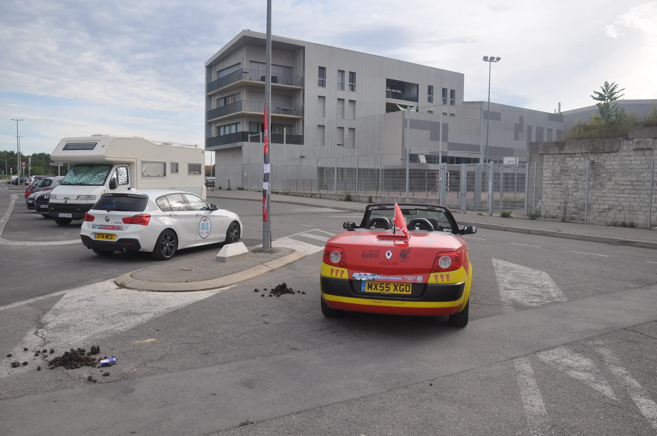 A small red and yellow children's toy car with Birmingham City Football Club decorations parked in a parking lot. Behind it, a white sports car with a taxi sign on the roof and a logo on the side. Further back, a larger white camper van, and several 