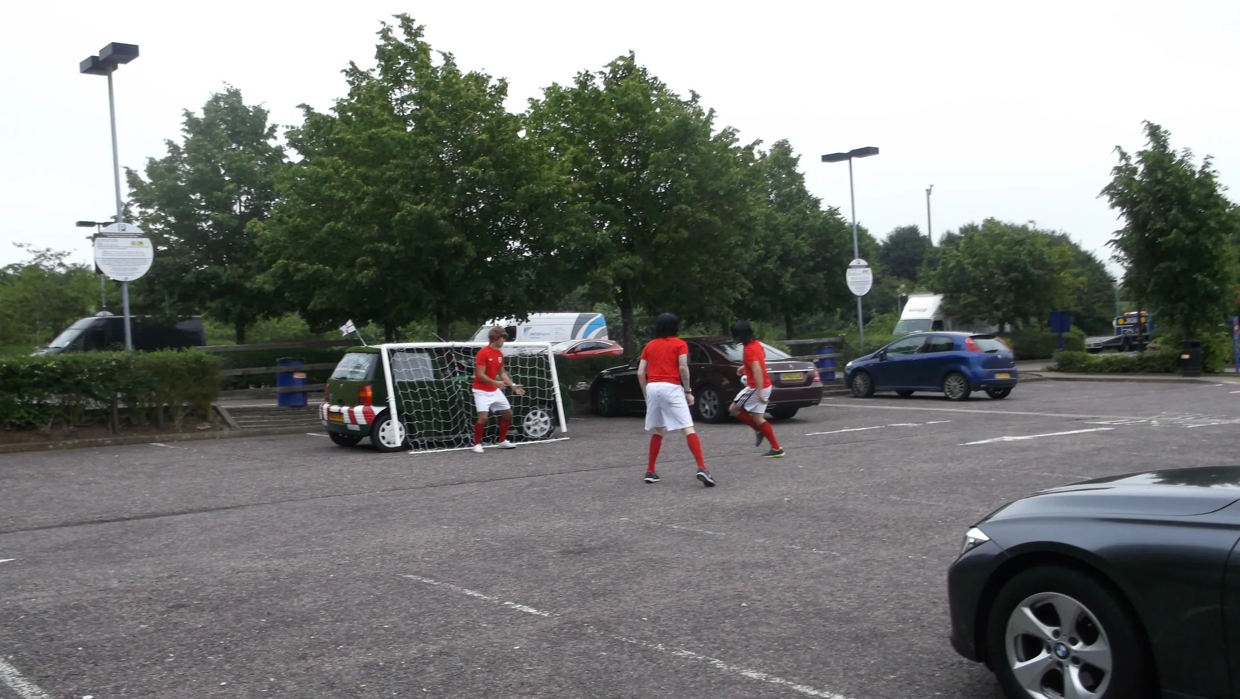 Three children in red shirts and white shorts playing soccer in a parking lot.