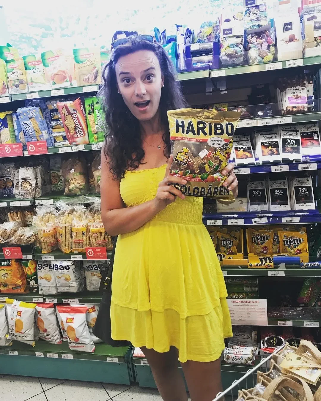 A woman with long, curly dark hair wearing a yellow dress stands in a grocery store aisle, holding a large bag of Haribo Gummy Bears and looking surprised.