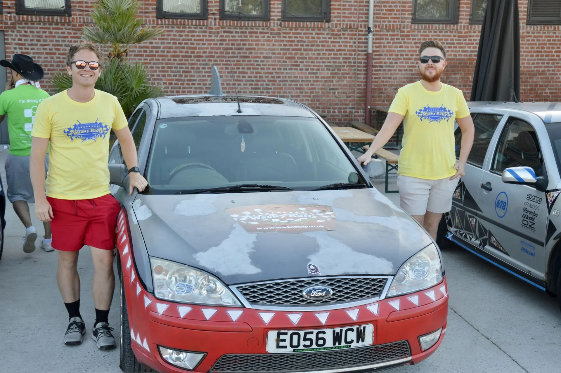 Two young men wearing yellow T-shirts with a race logo, standing next to a gray Ford car with red and white mountain-shaped decals on the bumper. The man on the left is smiling, wearing red shorts, black socks, and sneakers. The man on the right has 