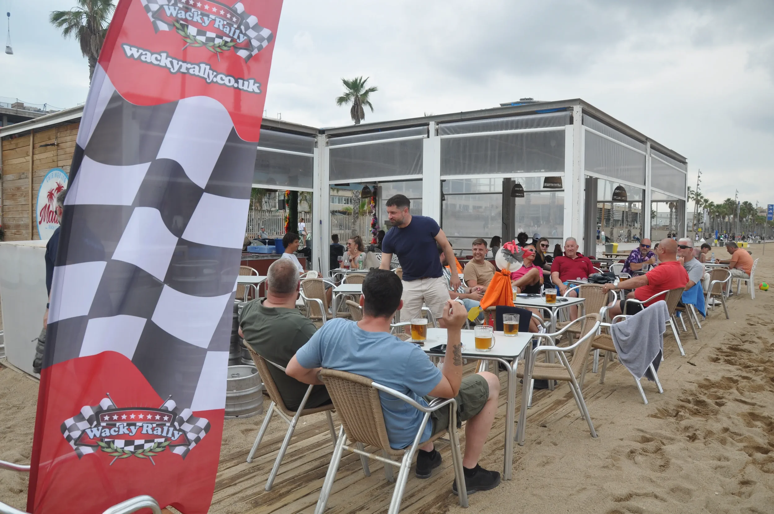 People seated at tables on the beach with a Wacky Rally flag in the foreground and a beachside structure in the background.