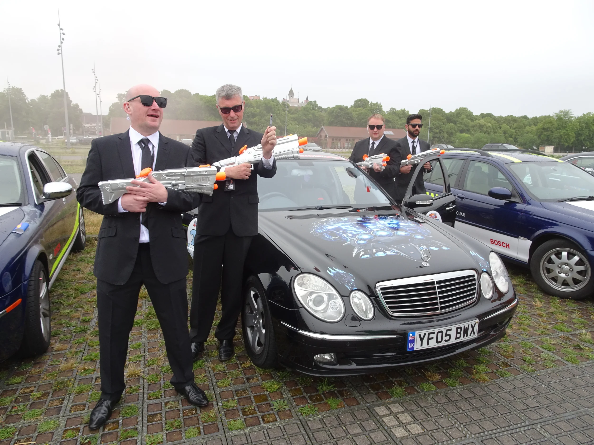 Five men dressed in black suits and sunglasses are holding toy water guns in a parking lot with police and security cars, a black Mercedes car with a holographic design on the hood, and a background of trees and a castle-like structure.