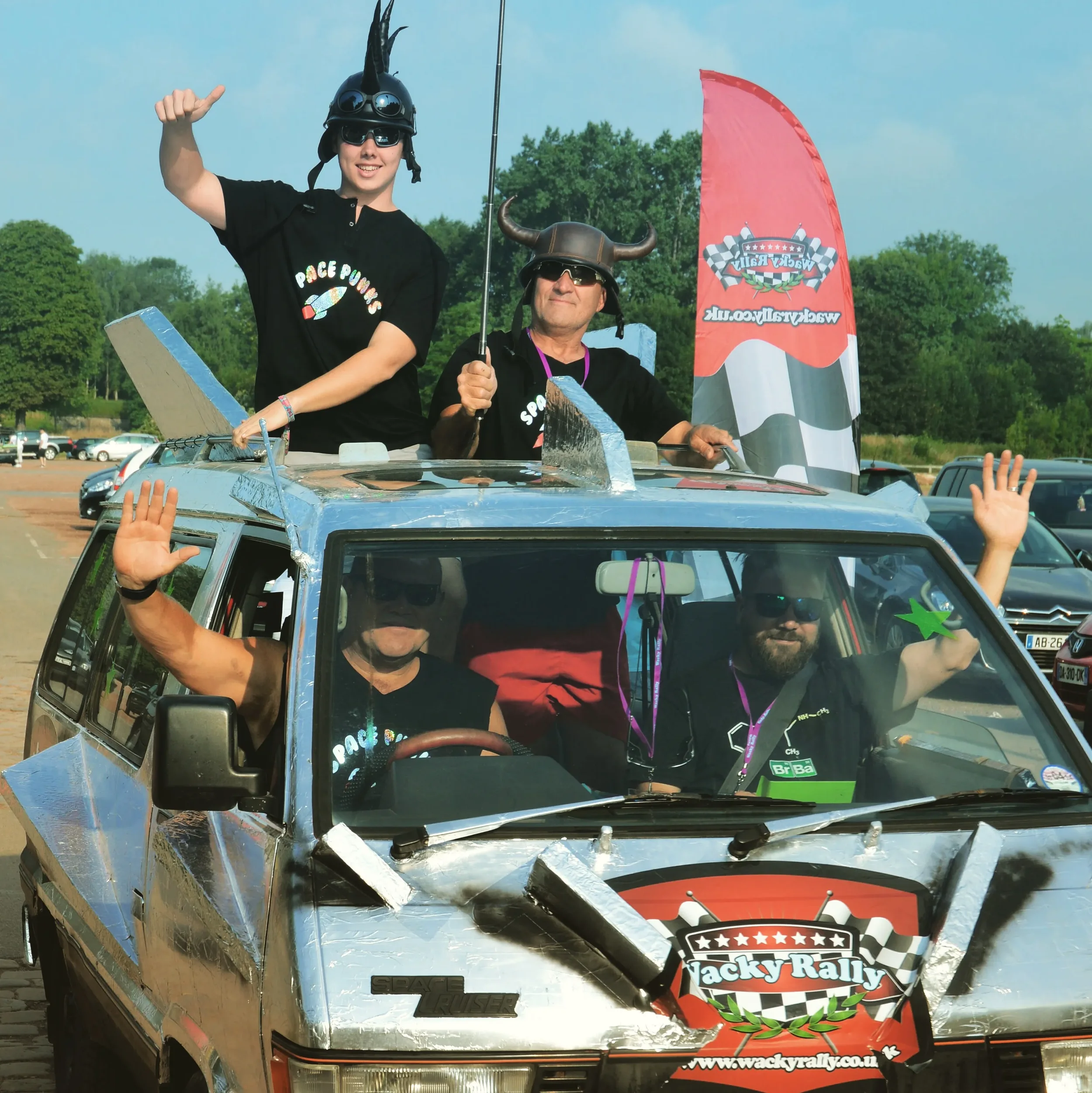 Four people enjoying a rally event on a decorated metal car, two are sitting in the car and two are standing on top, wearing helmets with horns, and smiling. The scene is set outdoors with a parking lot and trees in the background.