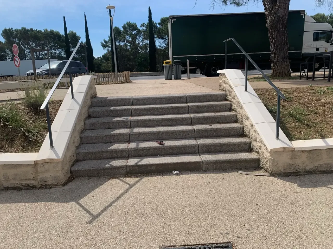 Concrete stairs with metal handrails leading up to a sidewalk in a park or outdoor area with trees and vehicles passing by.