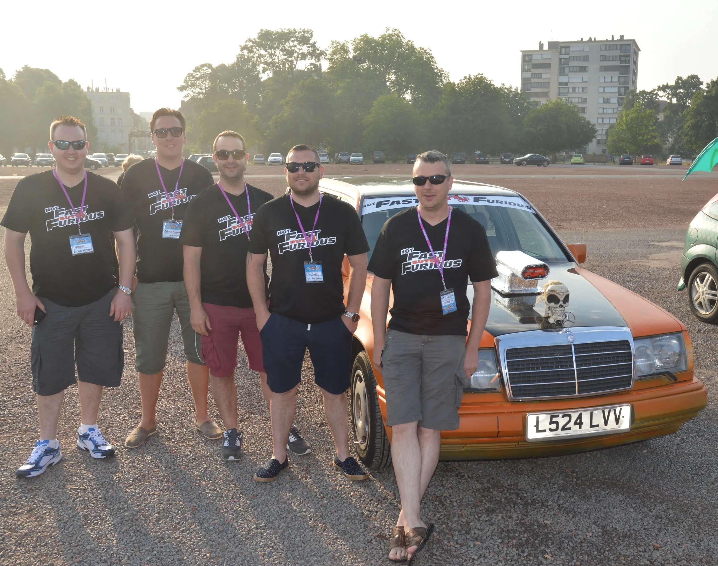 Five men standing in front of a vintage car with a skull and toy car on the hood, all wearing matching black 'Fast & Furious' t-shirts and lanyards, outdoors during daytime.
