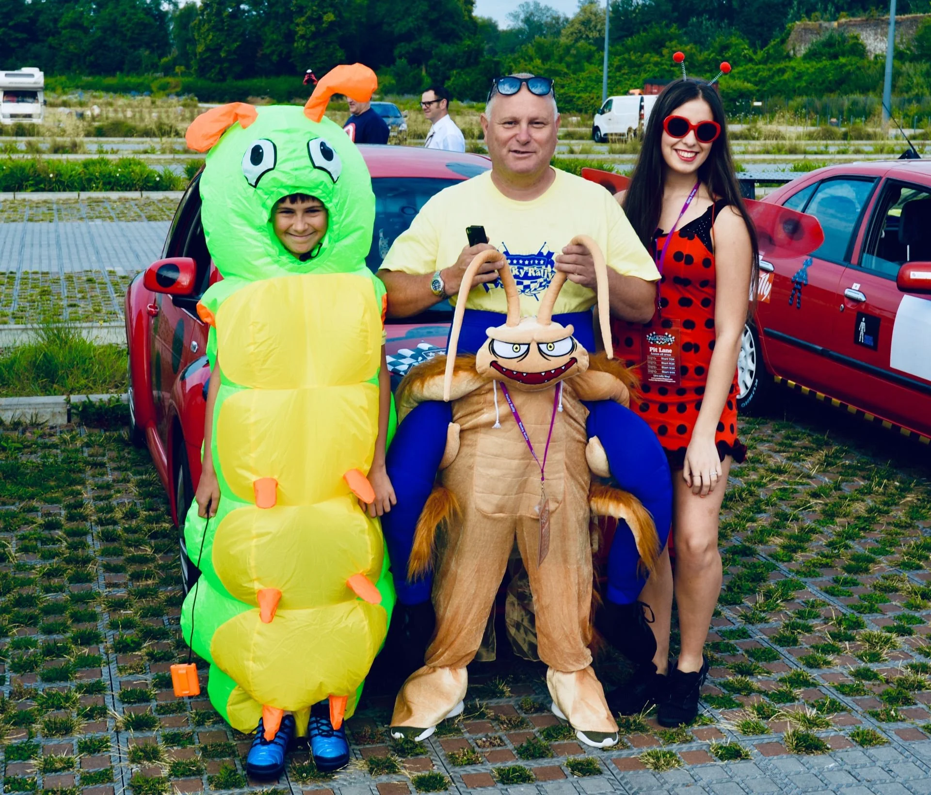 Three people dressed in costumes, standing outdoors near a red car, with other cars and people in the background. One person is dressed as a green caterpillar with an orange head, and there is a person in a crab costume with large claws. The third pe