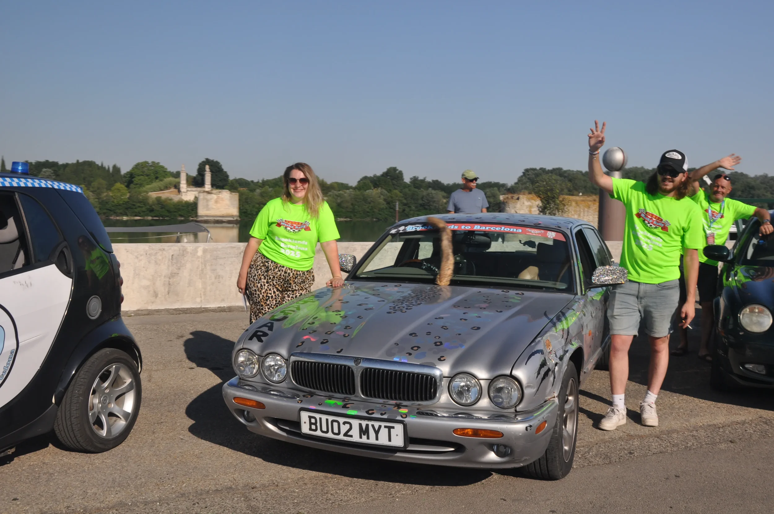 Group of people celebrating beside a decorated silver car on a bridge, with a woman in leopard print pants and green shirt, and a man with long hair and beard in bright green shirt making a peace sign, during a sunny day.