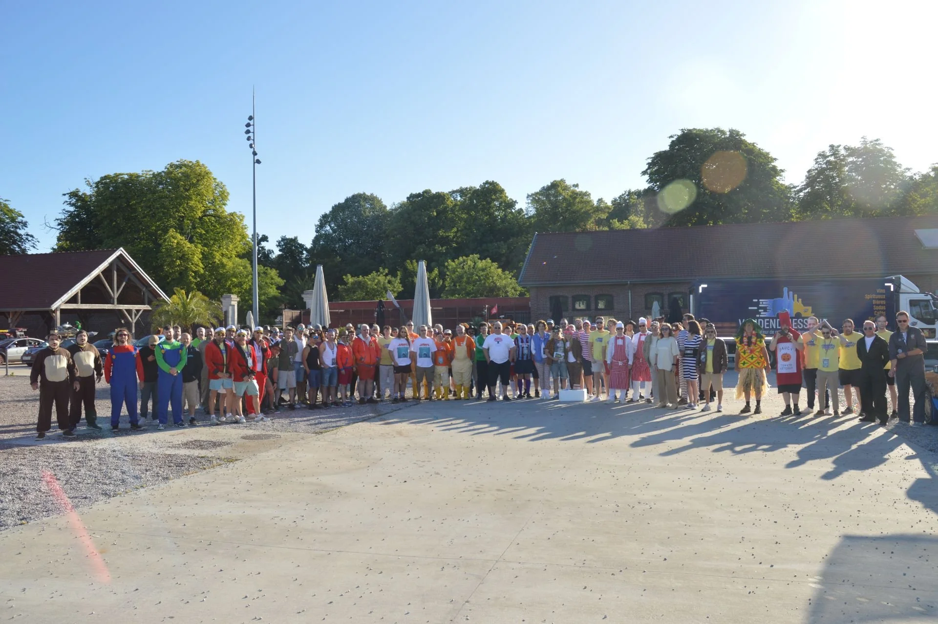 A large group of people dressed in various costumes, standing outdoors on a sunny day, with trees, buildings, and a truck in the background.