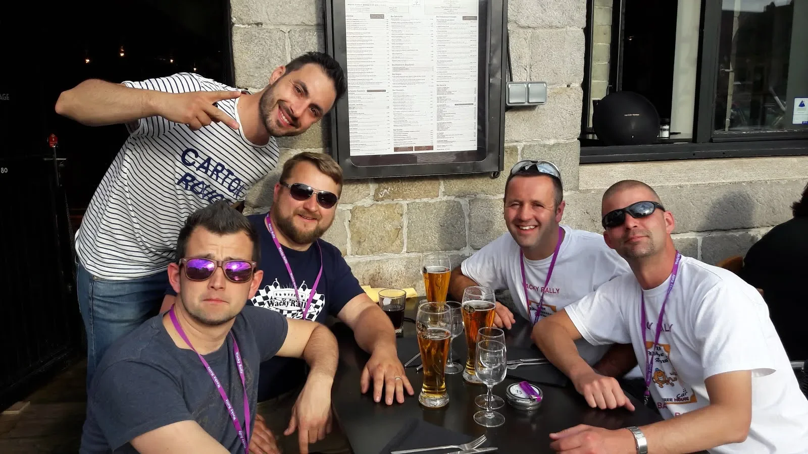 Five men sitting around a table with drinks outside a restaurant, four of them wearing sunglasses and purple lanyards, one man standing and pointing at the camera, all smiling.