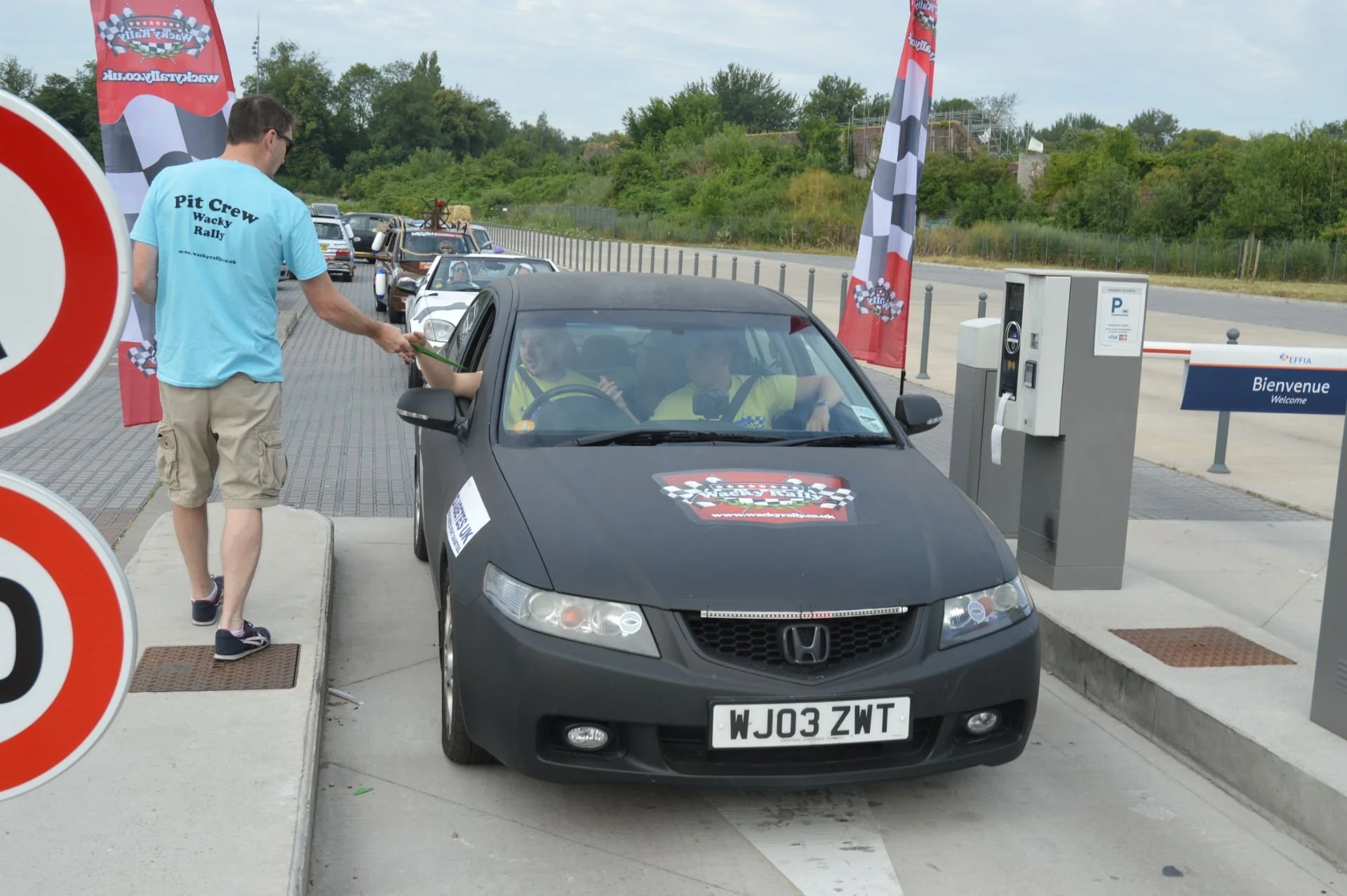 A black Honda car at a checkpoint with a driver and a passenger inside, interacting with a pit crew member during a rally event. There are flags with checkered patterns, a sign that says 'Bienvenue,' and other cars in line at a rally event.
