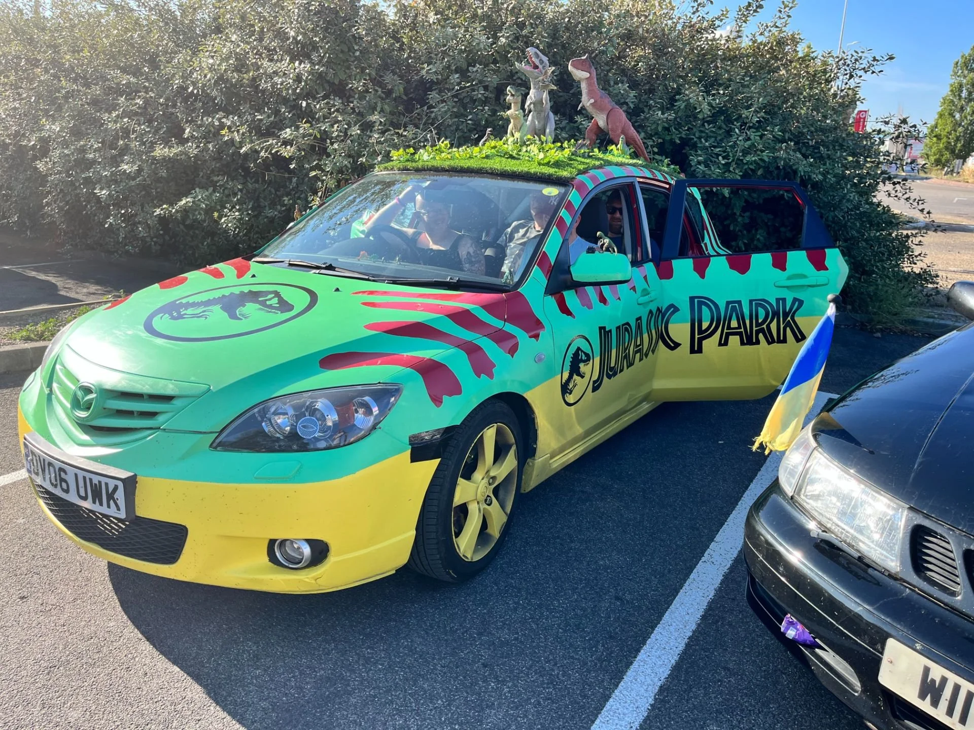 A colorful Mazda car with a Jurassic Park theme parked in a parking lot, decorated with a green and yellow paint job, red stripes, and a Jurassic Park logo on the hood. The car has dinosaur figures on the roof and an Ukrainian flag attached to the fr
