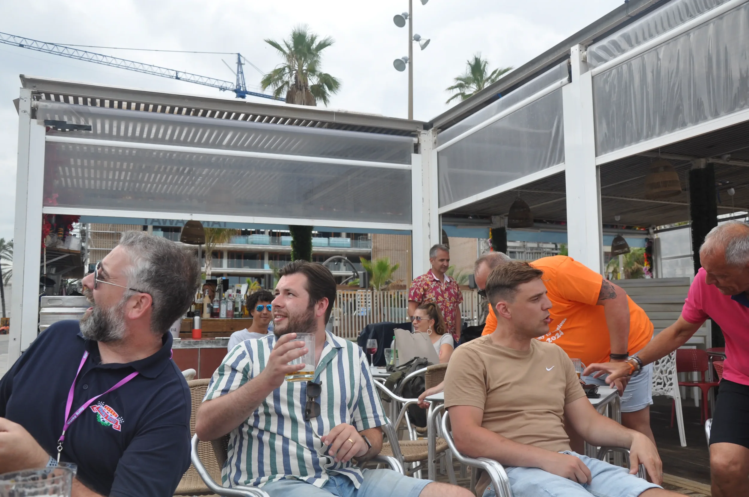 Group of people enjoying drinks at an outdoor bar or restaurant with palm trees, a crane, and buildings in the background.