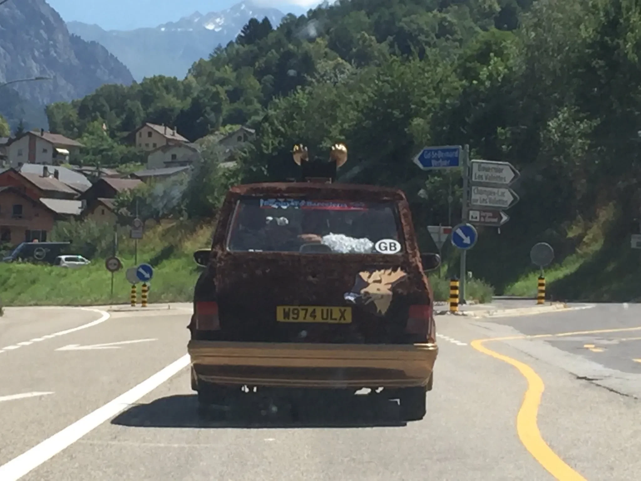 A small car with decorative reindeer antlers and a reindeer decal on the back is driving on a curved mountain road surrounded by trees and houses in a mountainous area.