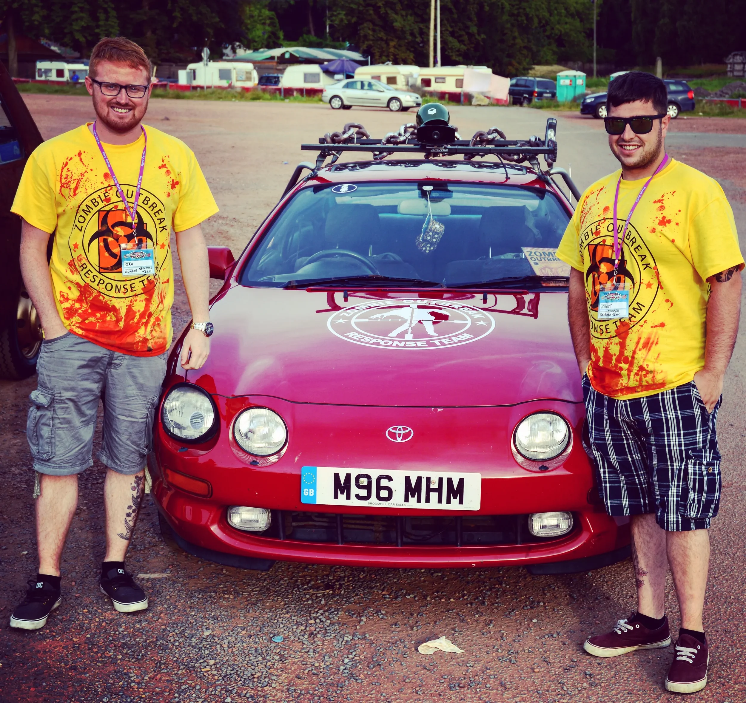Two men standing on either side of a red Toyota sports car in an outdoor parking area with camper trailers in the background. They are wearing yellow T-shirts with a logo and text, and both are smiling. The man on the left has glasses, khaki shorts, 