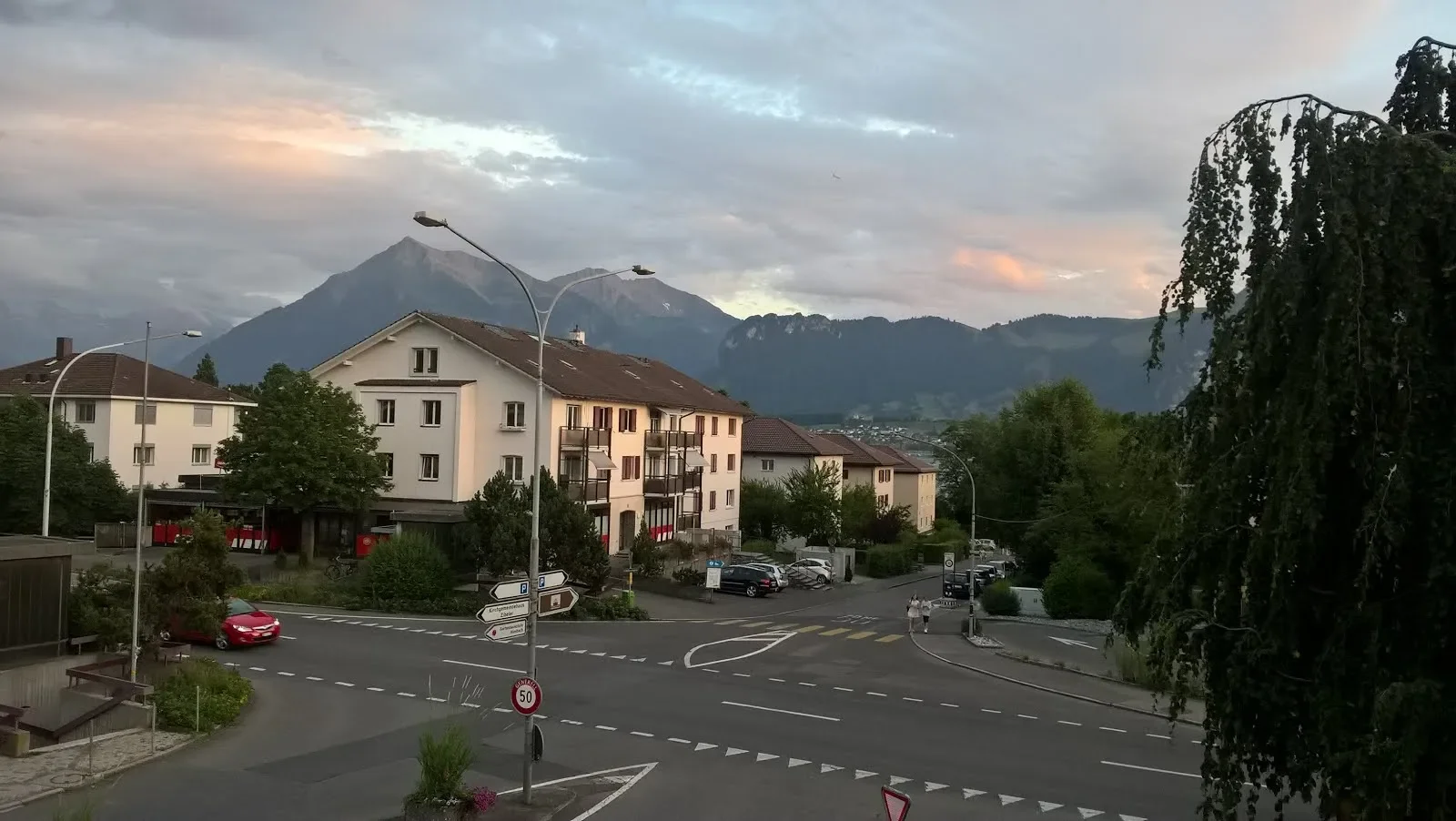 A street scene in a town with multi-story residential buildings, cars parked along the street, and mountains in the background under a partly cloudy sky during sunset.