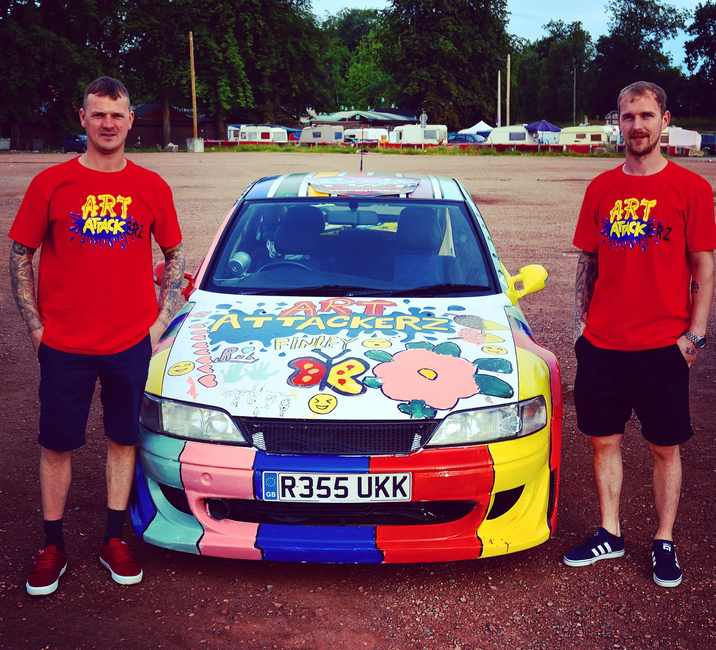 Two men in red t-shirts with a colorful art attack design stand beside a brightly painted car with similar artwork, in an outdoor setting with trees and tents in the background.