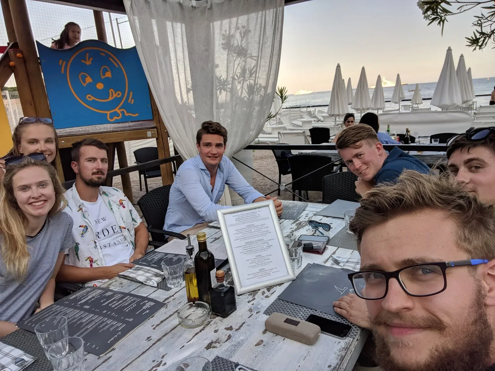 Group of young adults sitting at an outdoor restaurant table near the beach with white umbrellas and sailboats in the background, enjoying a meal.