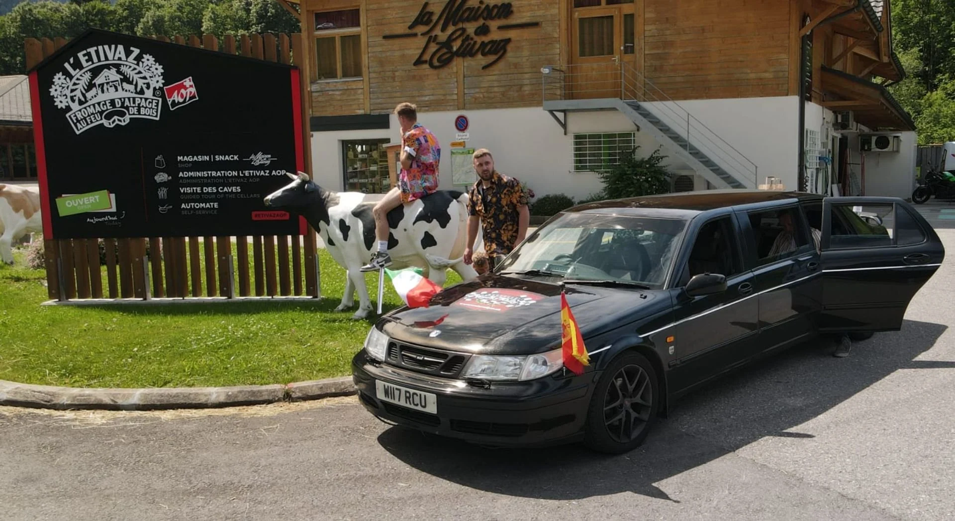 A black car parked on the street with a Spanish flag attached to the front and a second flag on the hood, in front of a wooden building. Two men, one sitting on a life-sized cow statue and the other standing beside it, are dressed in colorful, patter