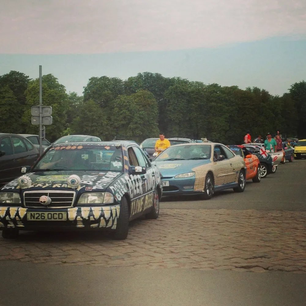 Line of cars decorated with various creative and colorful designs parked on a brick-paved area with trees and a cloudy sky in the background.